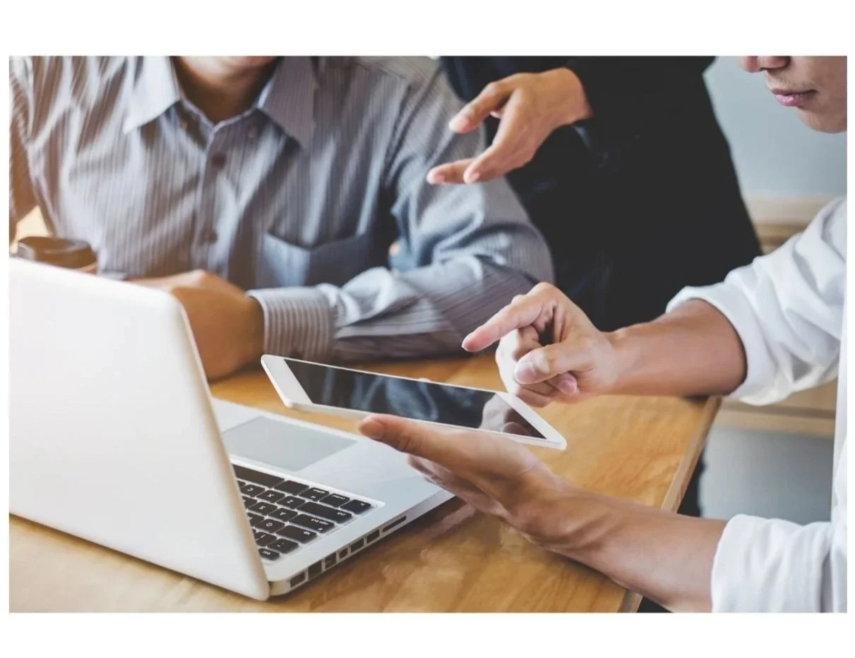 Two people are having a discussion while looking at a tablet and laptop on a wooden table.