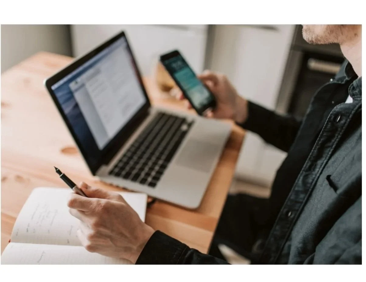 Person sitting at a wooden table with a laptop, a smartphone, and a notebook, writing in the notebook while looking at the phone.
