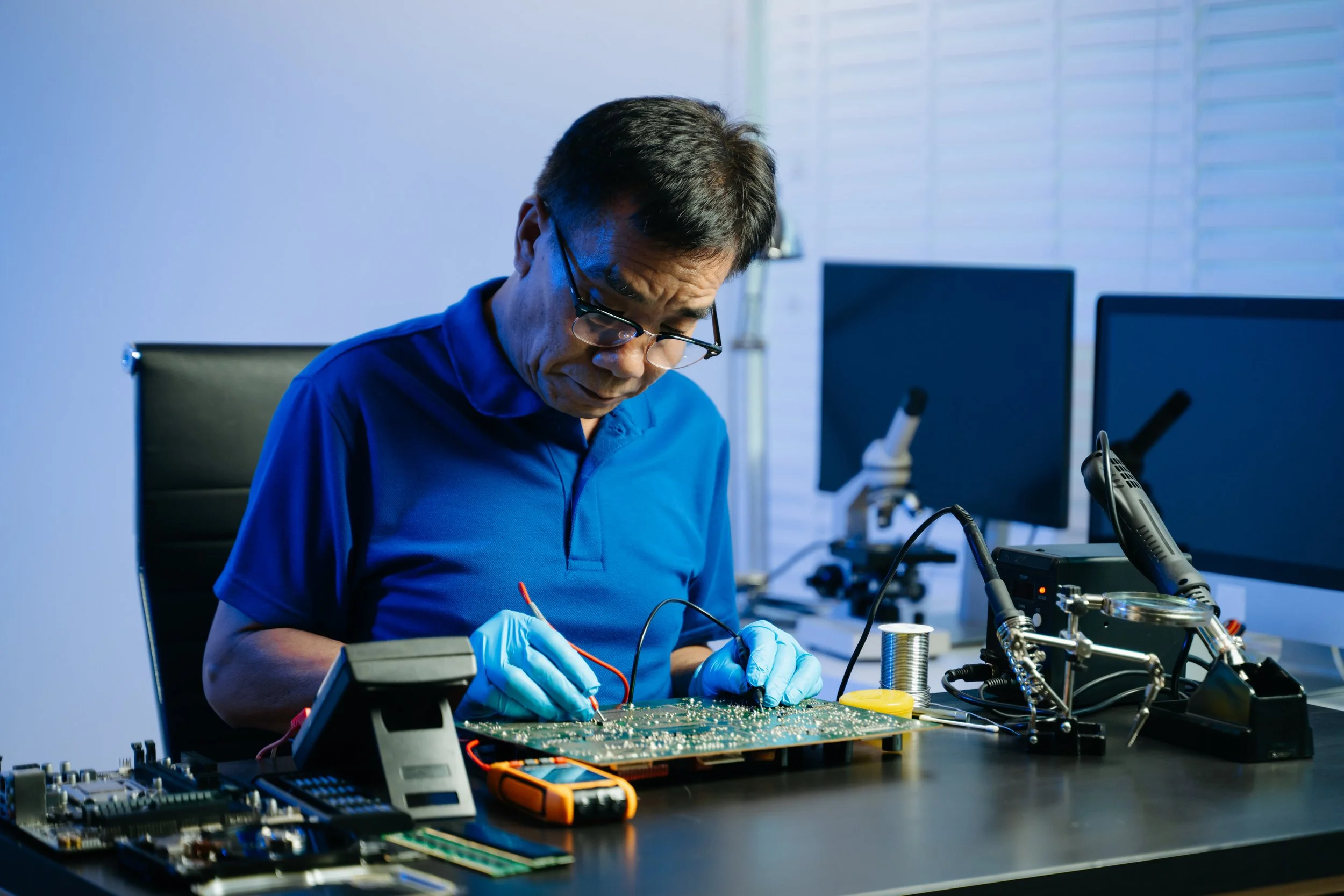 A man wearing glasses, blue shirt, and blue gloves working on an electronic circuit board in a high-tech laboratory with microscopes, monitors, and electronic equipment.