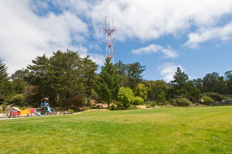 A park with a playground, trees, and a tall radio tower in the background under a partly cloudy sky.