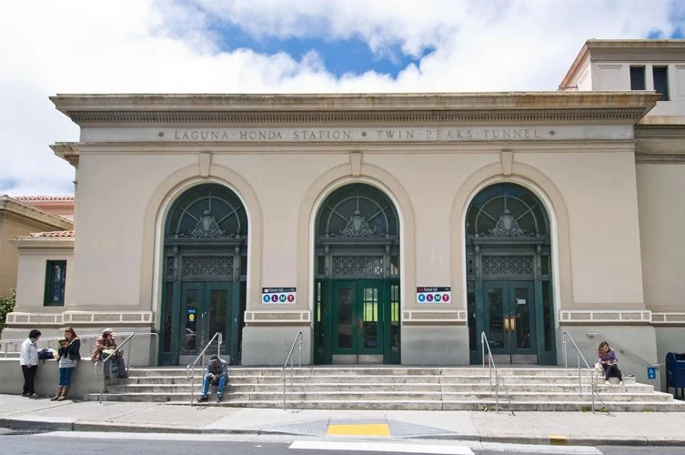 Exterior of the Laguna Honda Station with three tall arched windows and steps leading up to the entrance, people sitting and standing outside.