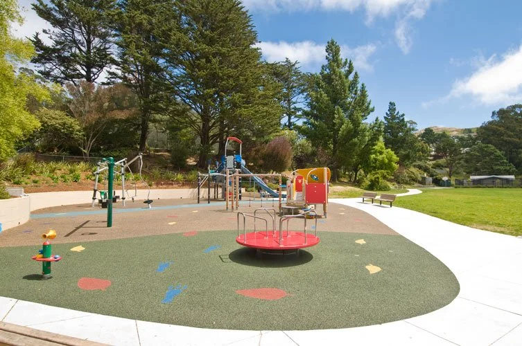 Empty playground with a merry-go-round, swings, slides, and a spring rider in a park surrounded by trees and a grassy area under a partly cloudy sky.