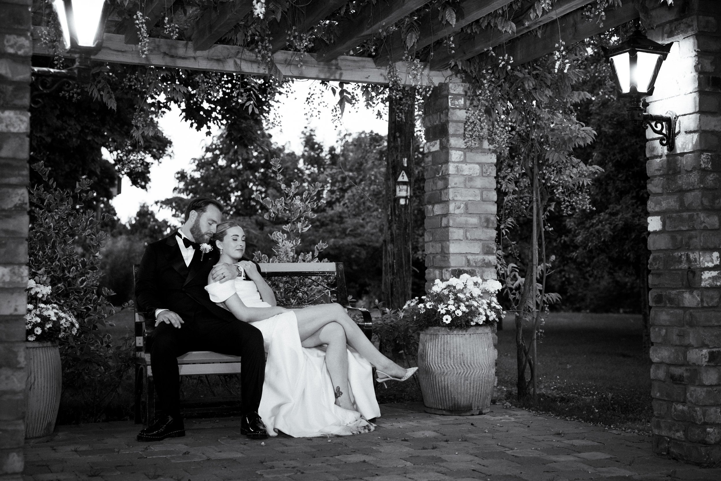 A black and white photo of a couple sitting on a bench under a pergola adorned with hanging plants and flowers. The man is dressed in a tuxedo, and the woman is in a white dress, with one leg crossed over the other, showing a tattoo on her ankle.