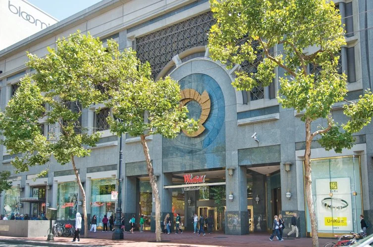 People walking in front of a shopping mall with green trees and a decorative circular design on the building facade.