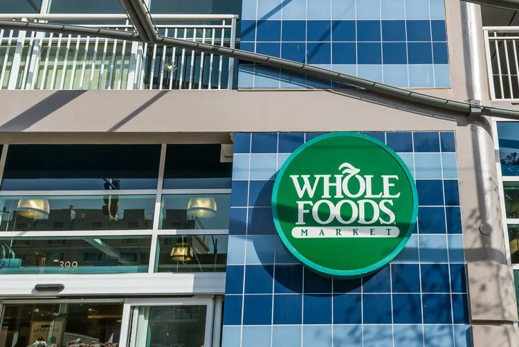 Exterior of a Whole Foods Market store with a large round green sign featuring the Whole Foods Market logo on a blue tiled wall.