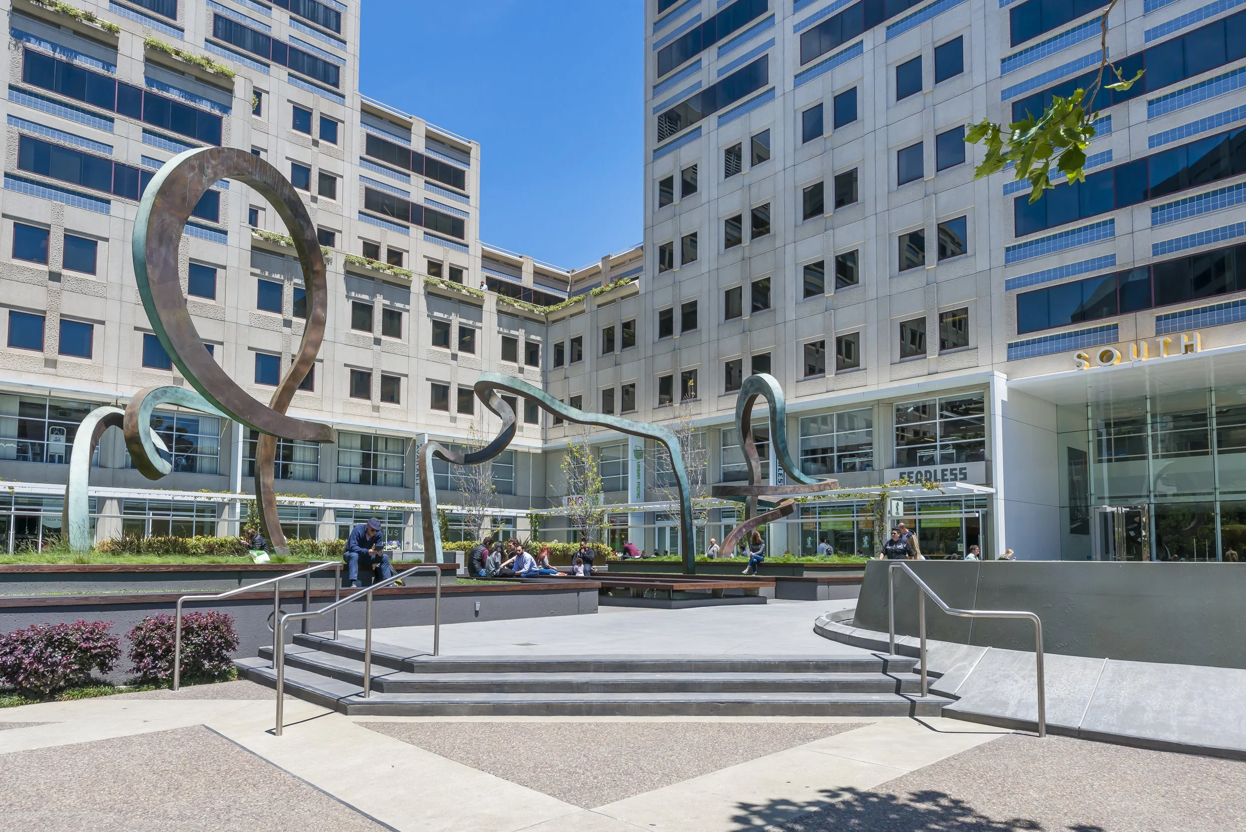 Urban outdoor plaza with modern sculptures, people sitting, and tall commercial buildings under a blue sky.