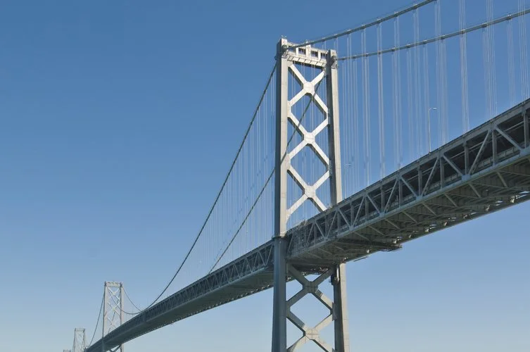 Close-up view of a large suspension bridge against a clear blue sky, showing the bridge tower and cables.