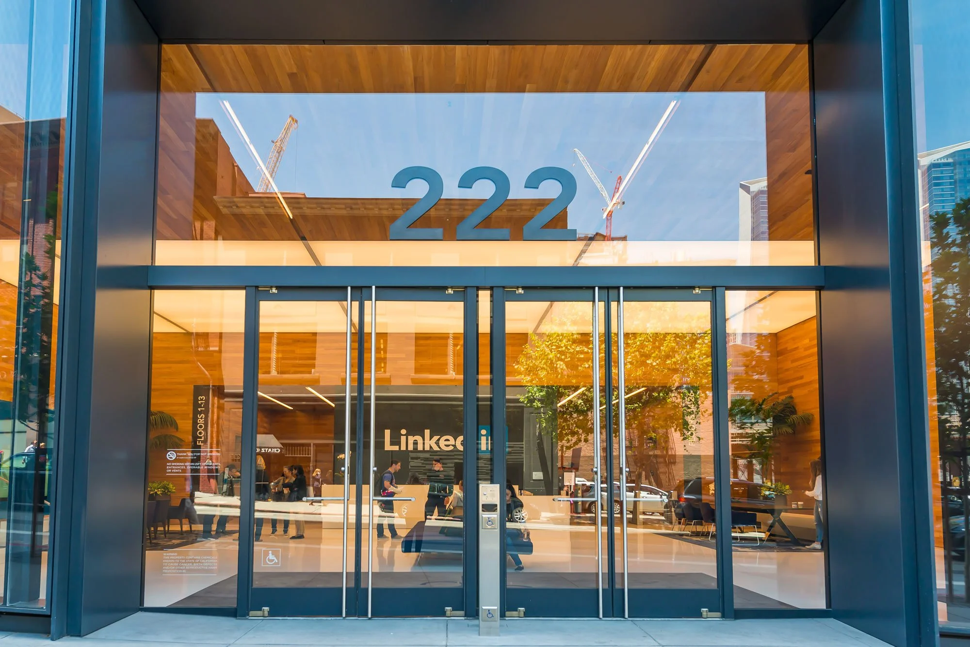 Modern building entrance with large glass doors, the number 222 displayed above, and the interior visible through the glass, showing people inside and trees reflected in the glass.