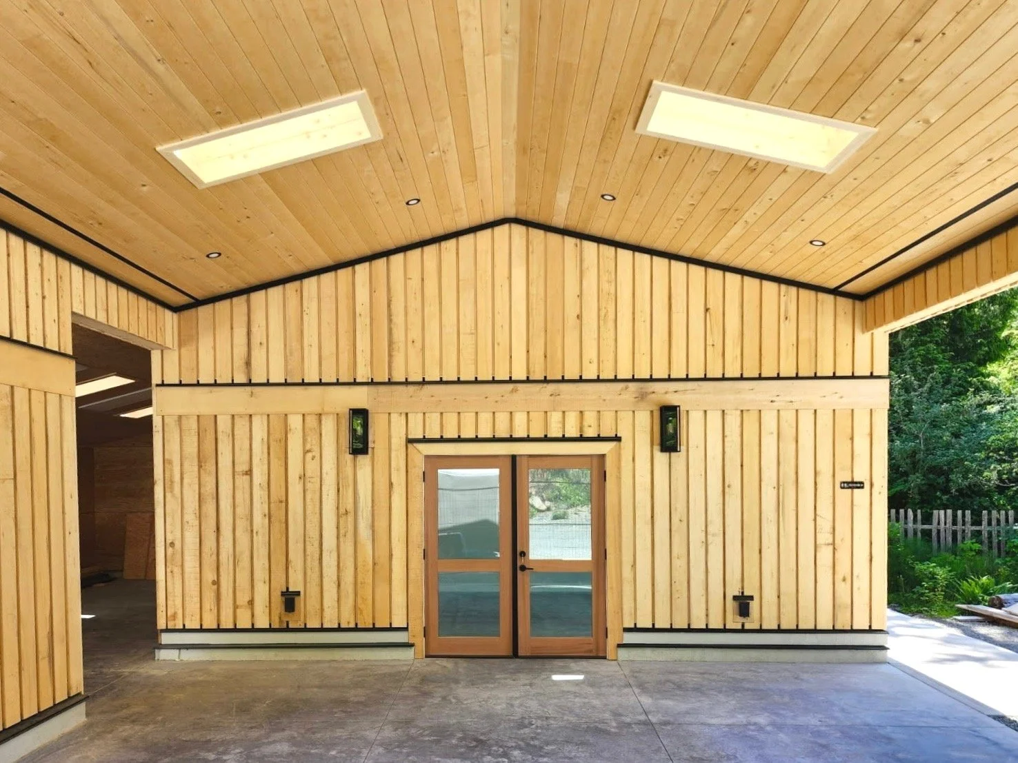 Exterior of a building with wooden walls and ceiling, a set of glass double doors, and outdoor greenery visible through the open area.