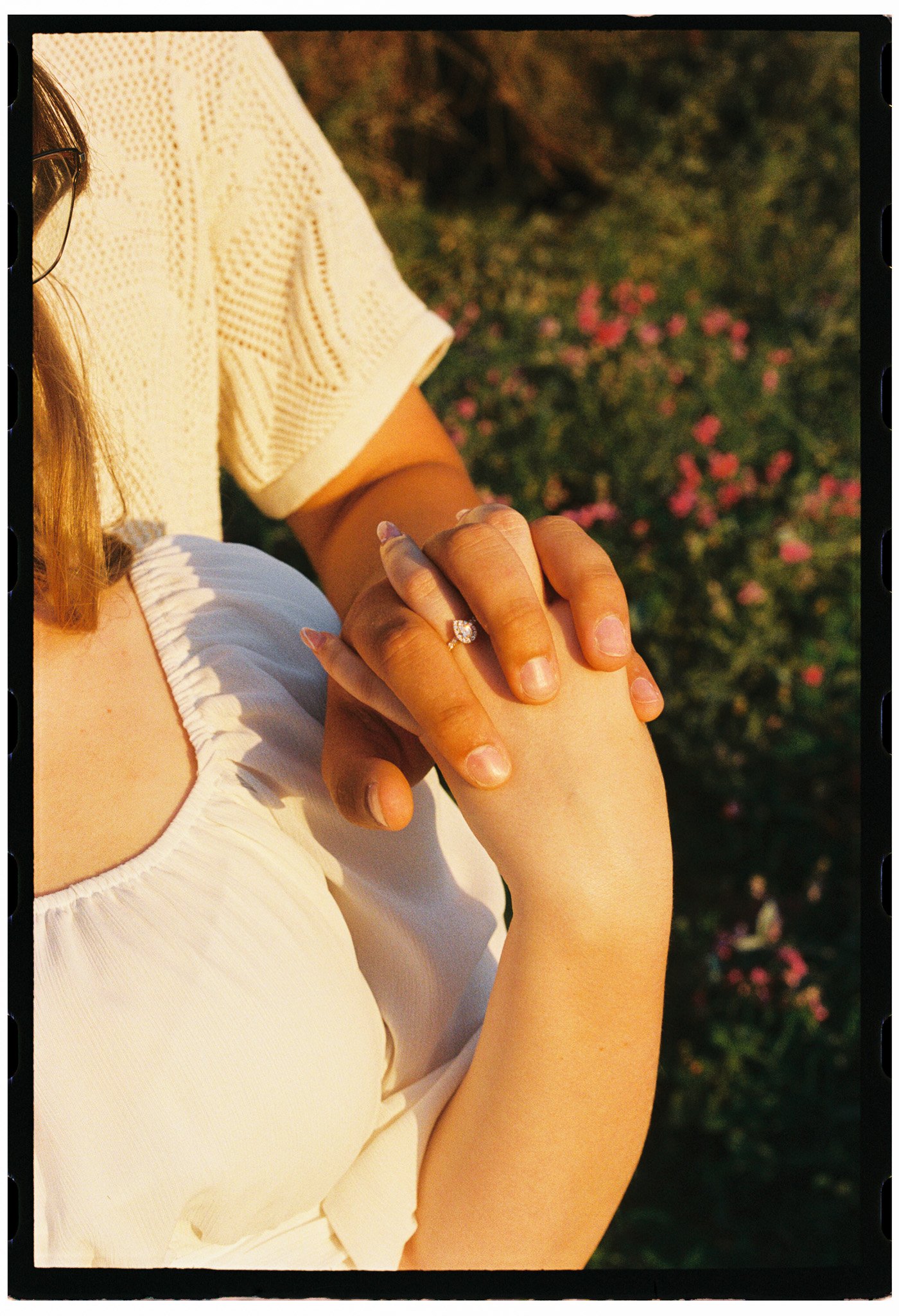 Discovery Park Close-up of partners holding hands, one with an engagement ring, outdoors with pink flowers in the background.