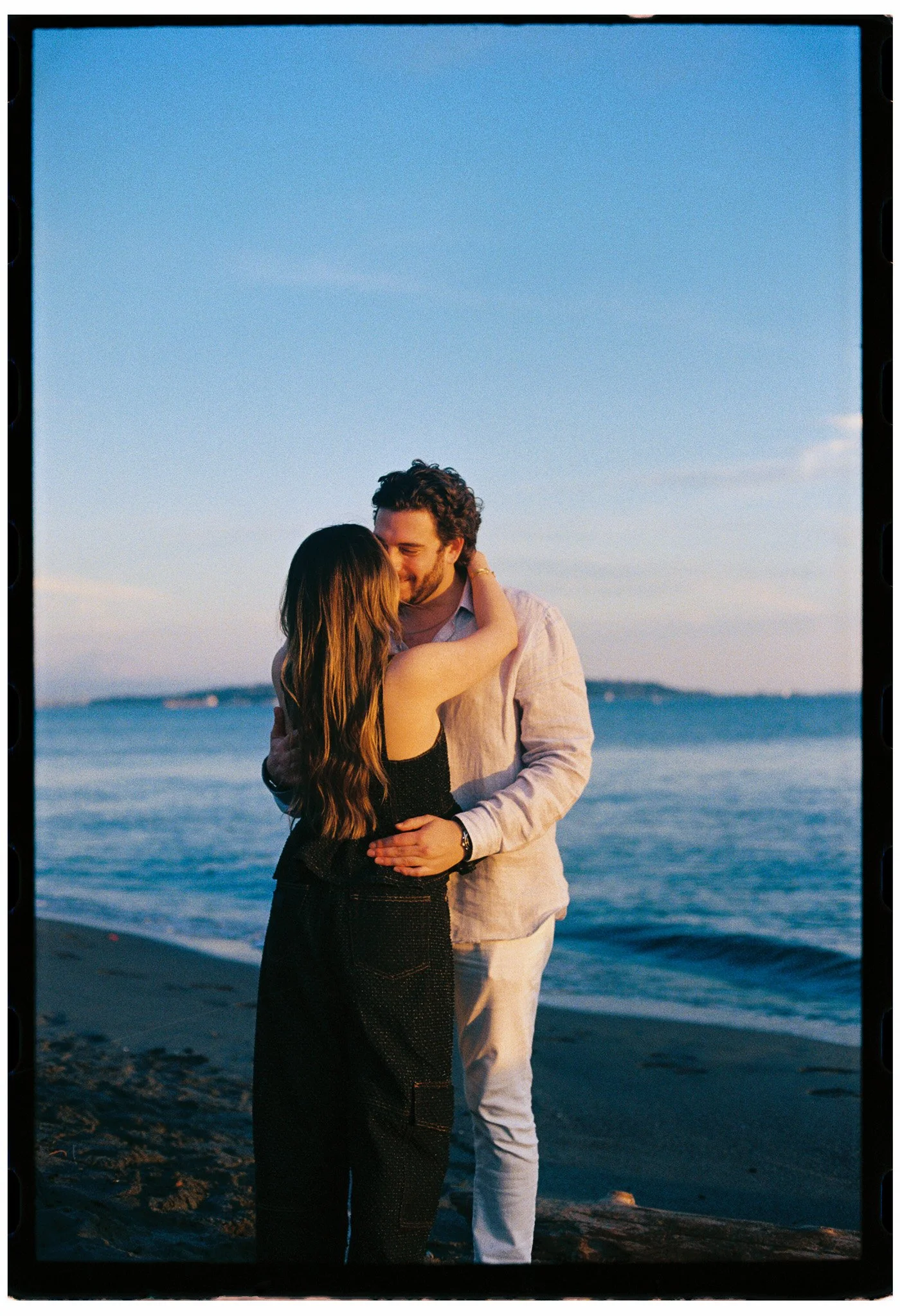 A couple hugging and smiling on a beach at sunset, with ocean waves and a distant shoreline in the background.