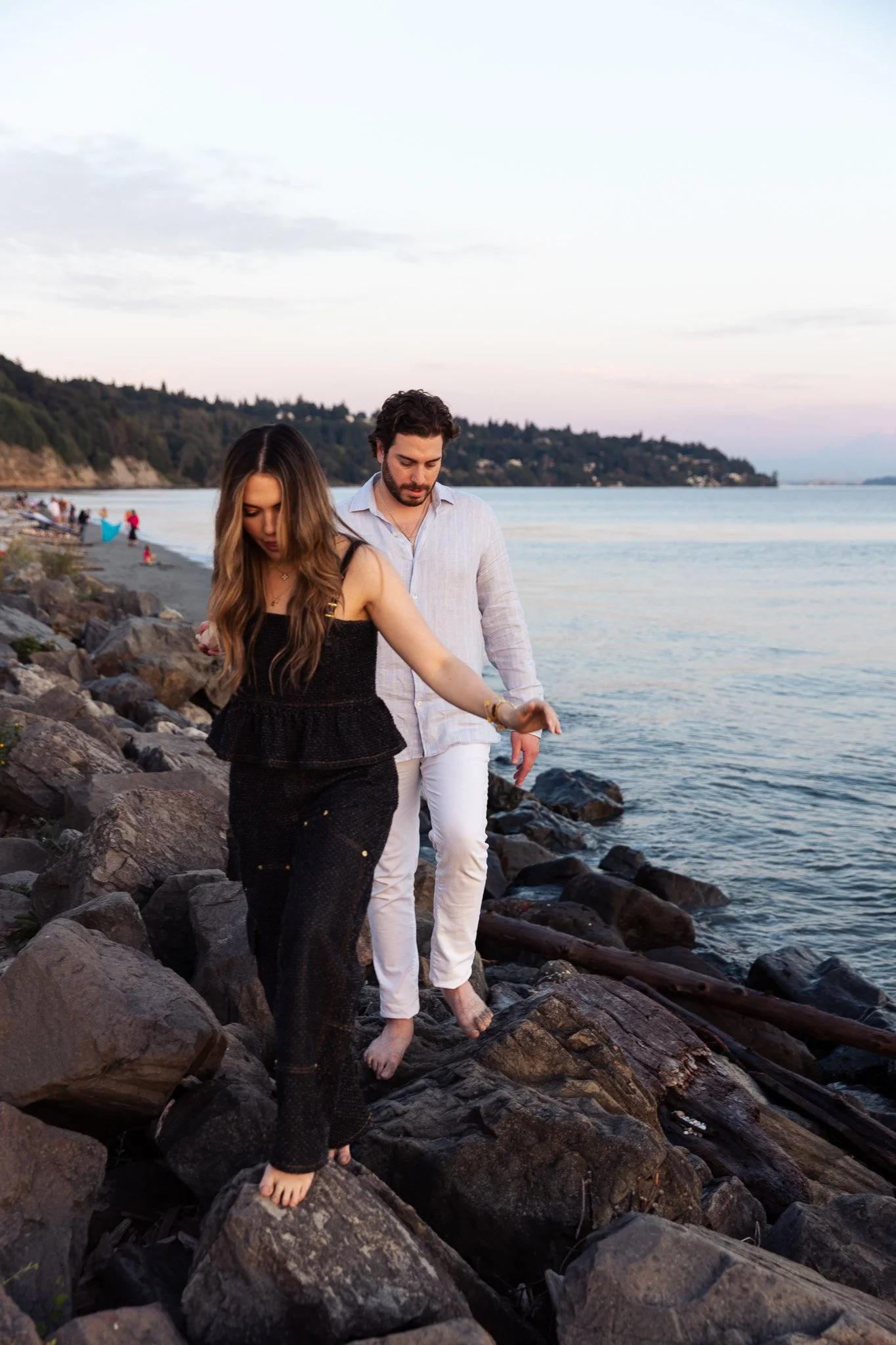 West Point Lighthouse Discovery Park Engagement in Seattle, Washington, partners walking barefoot on rocks along a beach with water and hills in the background during sunset.
