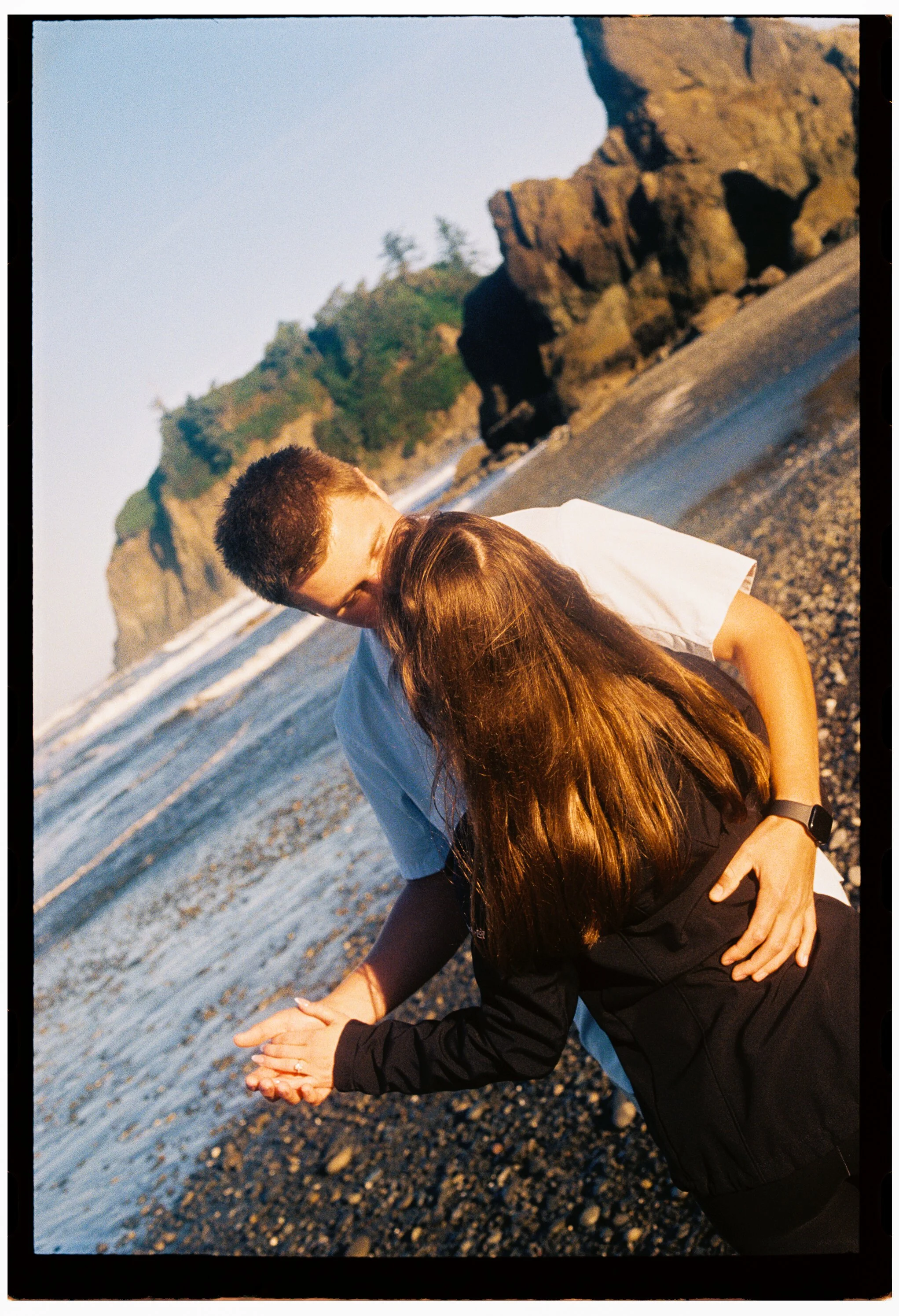 A couple sharing a kiss on a rocky beach at sunset, with cliffs and trees in the background.