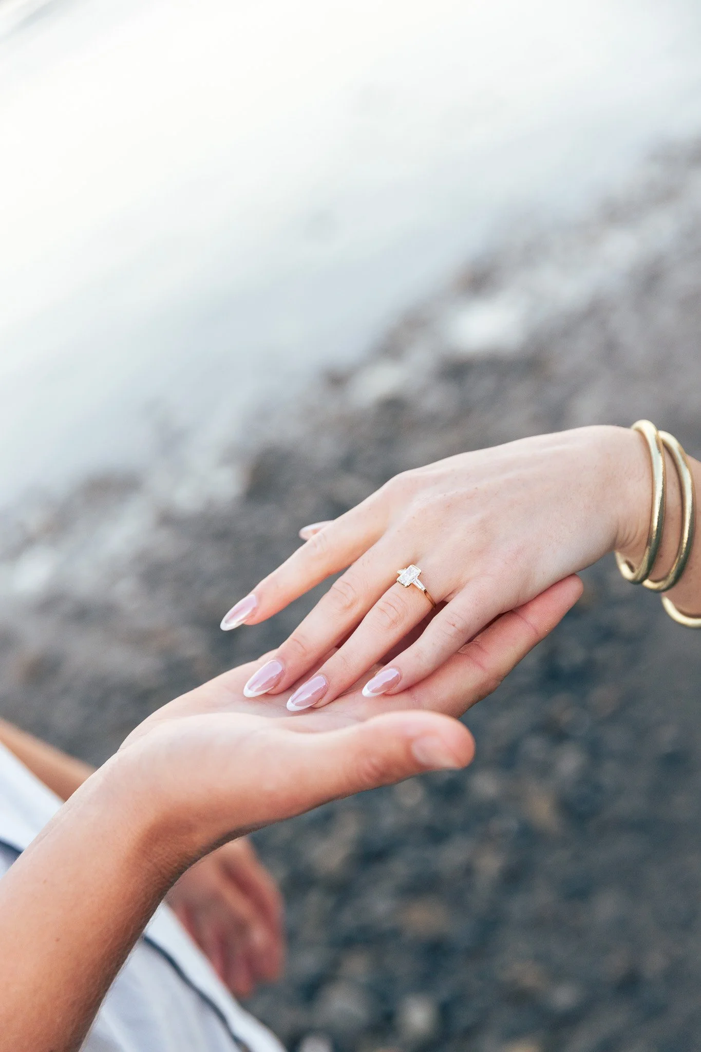Close-up of two hands gently touching, with one wearing a diamond ring and gold bangles, against a blurred background of water or beach.