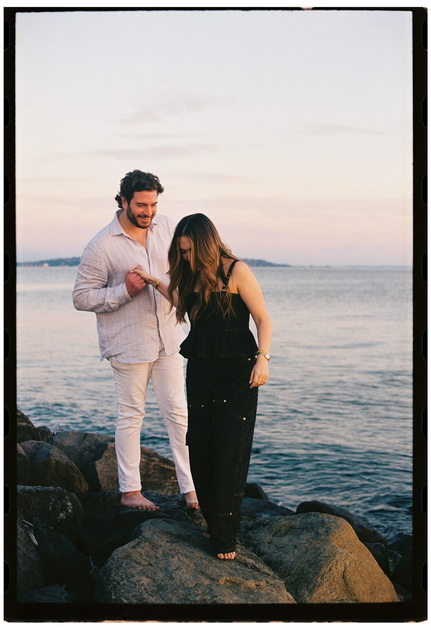 A man and woman standing on rocks at the beach during sunset, smiling and holding hands.