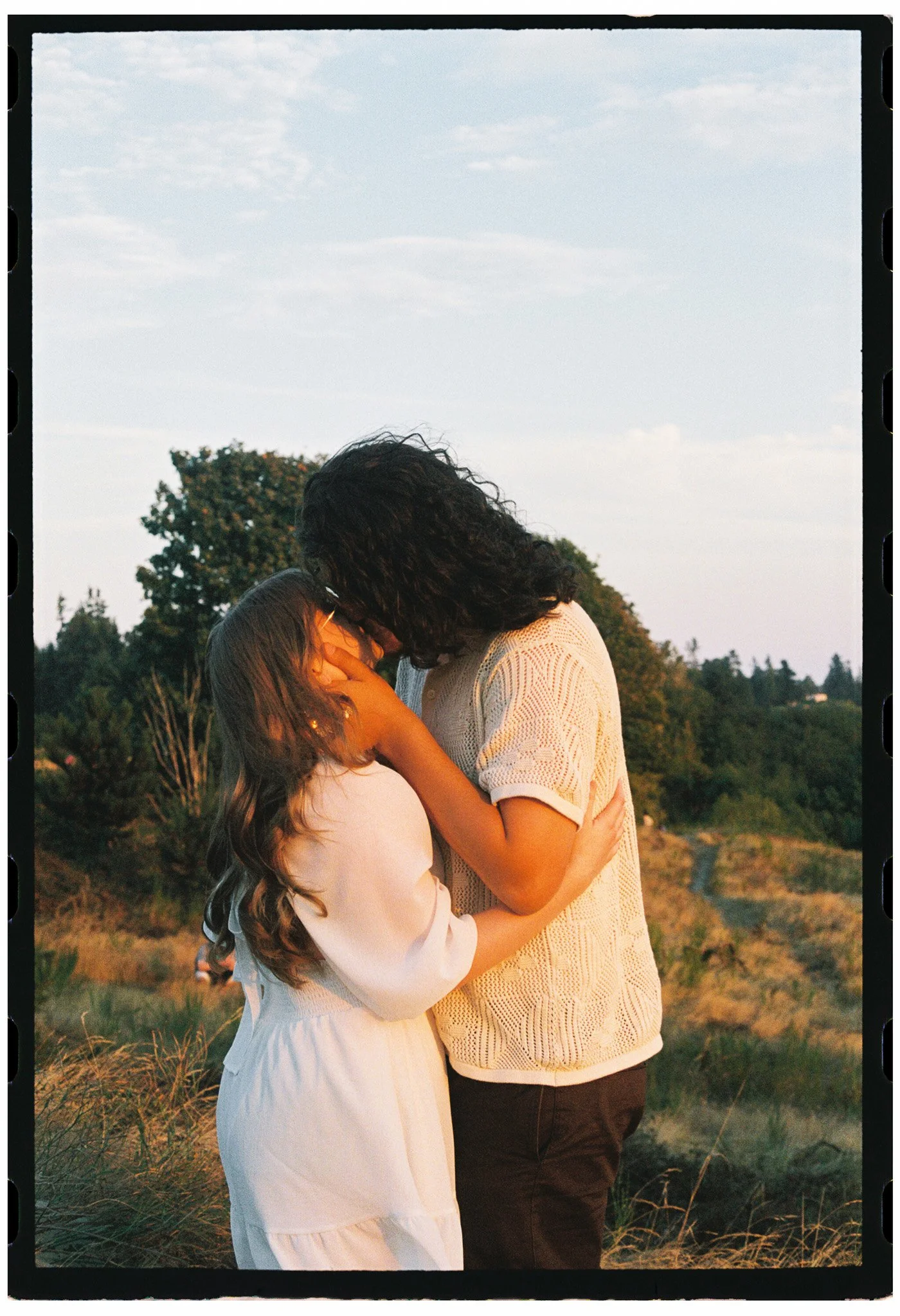 A couple lovingly embraces outdoors during sunset at Discovery Park, against a rural landscape background with trees and a cloudy sky.