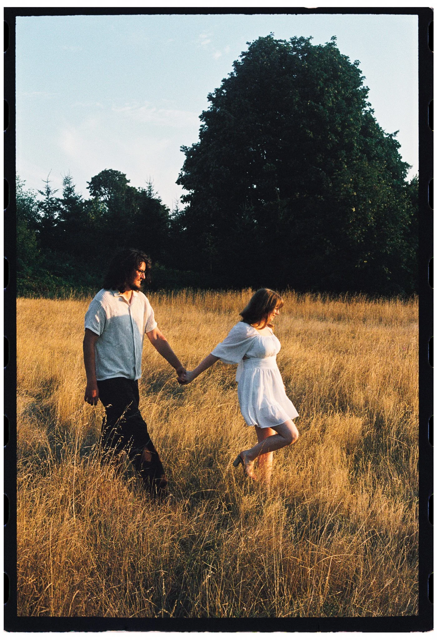 A man and a woman holding hands and walking through a field of tall grass during sunset for an engagement shoot at discovery park
