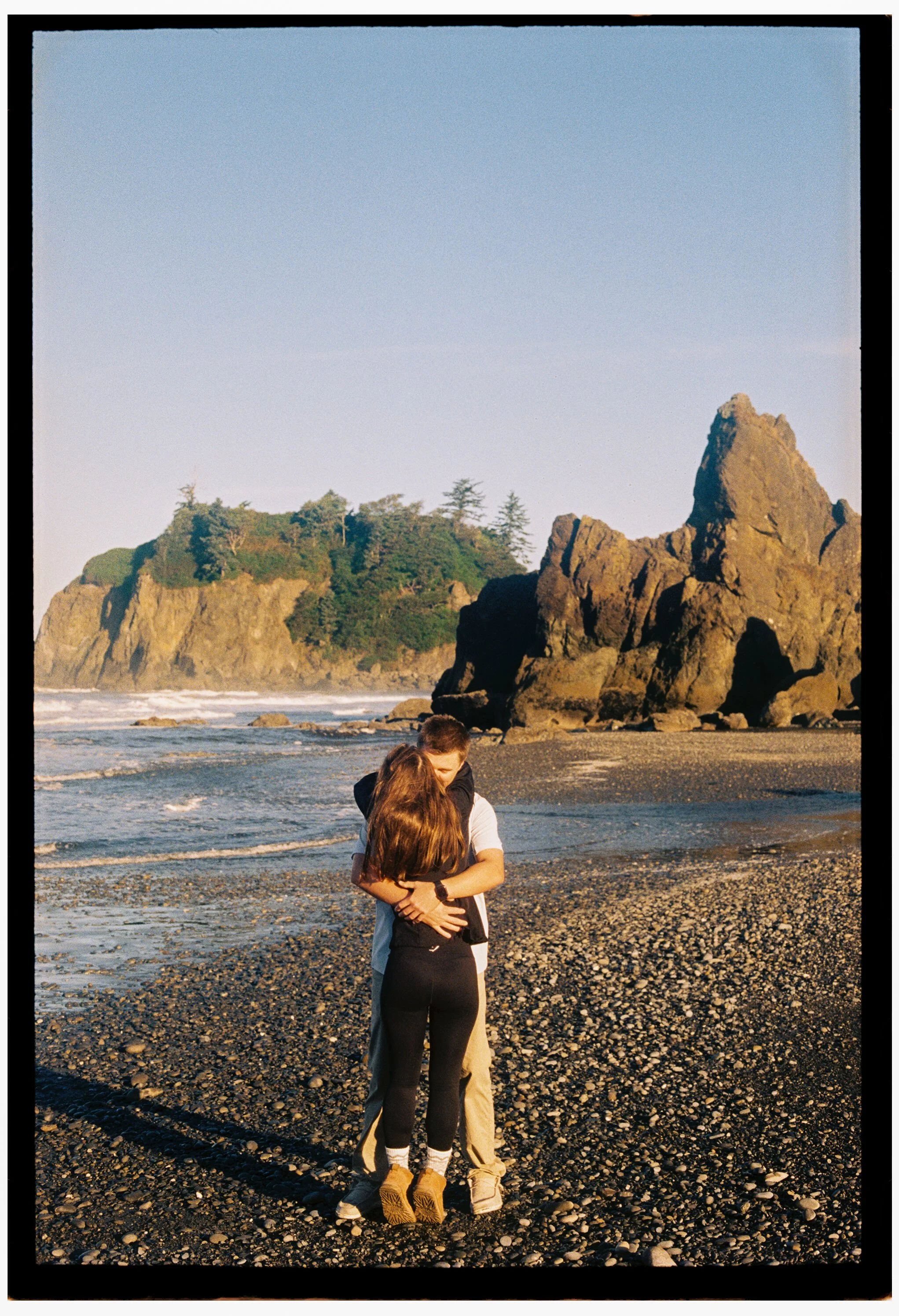 An engagement couple hugging on a rocky beach during sunset with large rocky formations and green trees at Ruby Beach, Washington