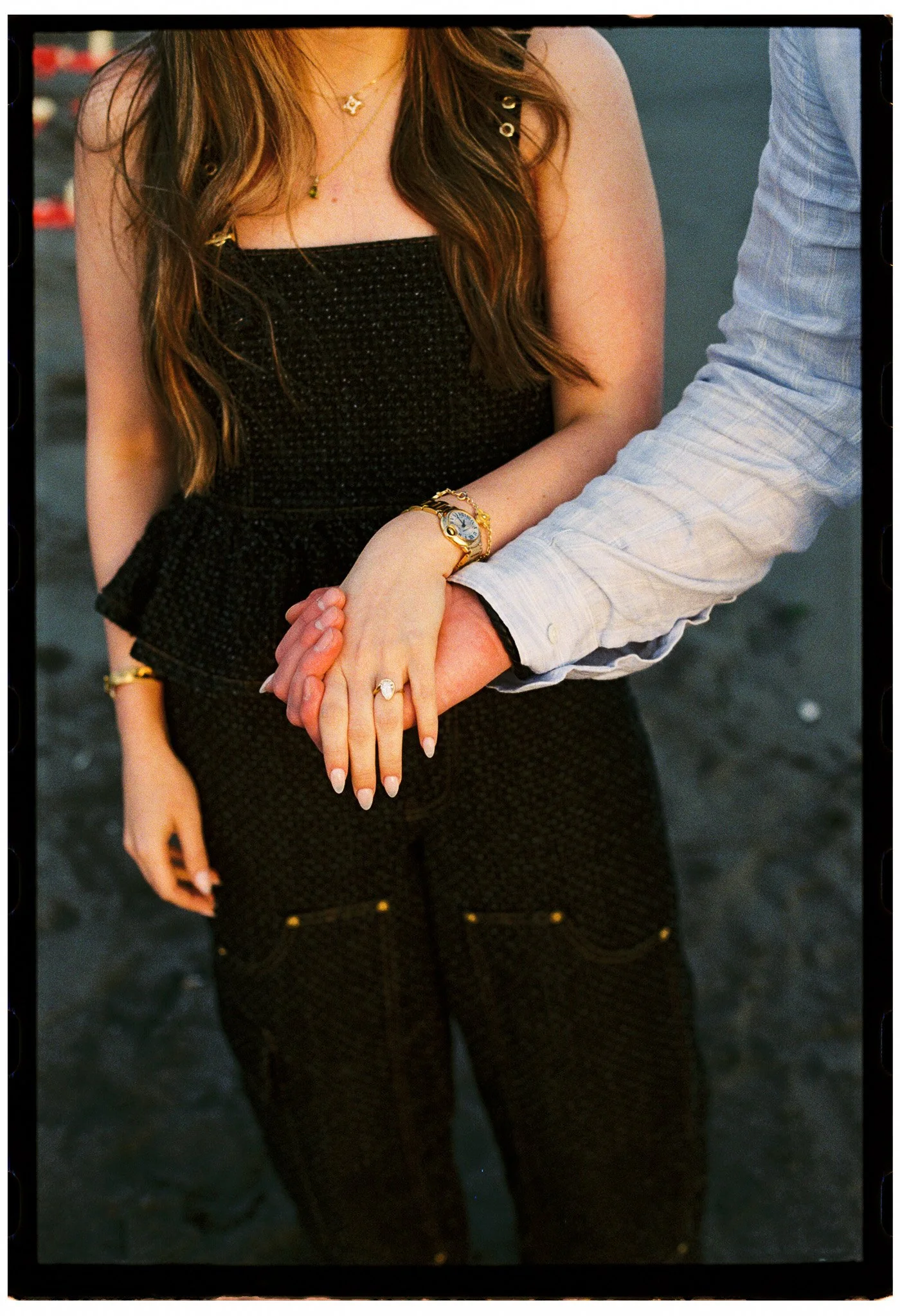 A woman and a man holding hands, her wearing a black dress with gold jewelry, including a watch and rings, and the man dressed in a light blue button-up shirt.