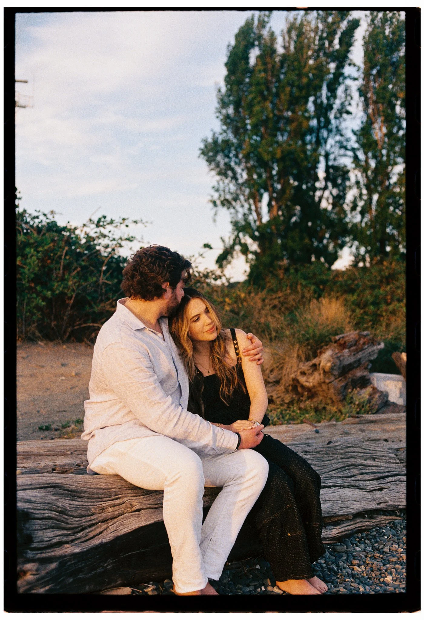 engagement couple sitting on a large piece of driftwood by the beach, hugging and smiling, with trees and a cloudy sky in the background during sunset at Discovery Park in Seattle, Washington
