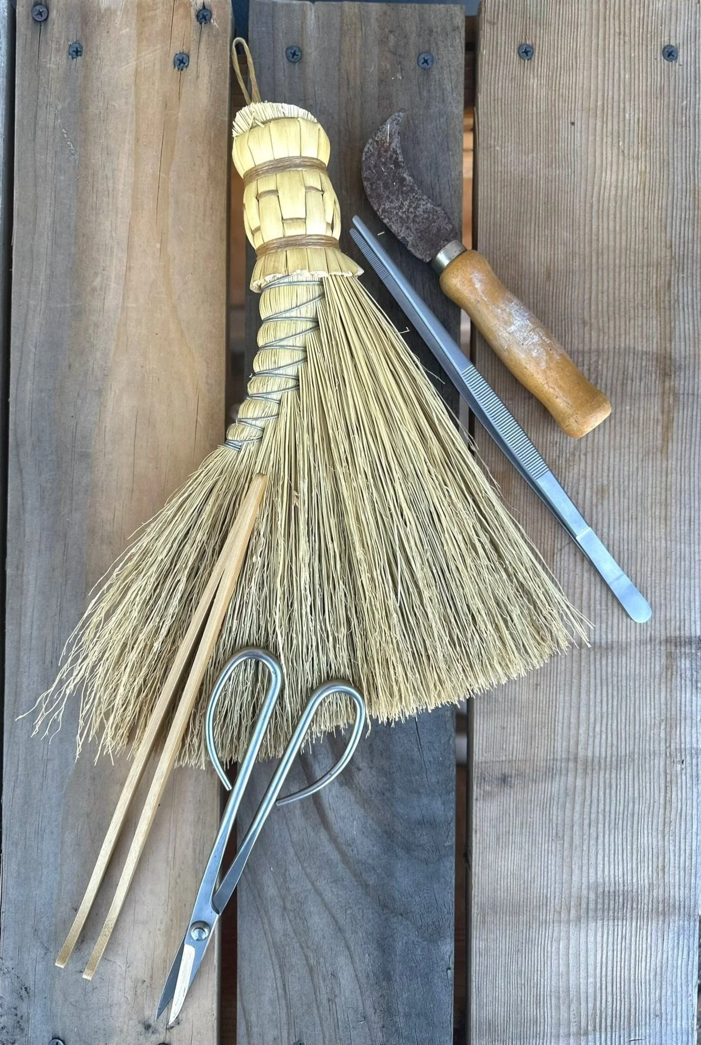 A small straw broom with a wooden handle, two wooden sticks, a metal pair of scissors, a metal comb, and a small curved sickle with a wooden handle are placed on a wooden surface.