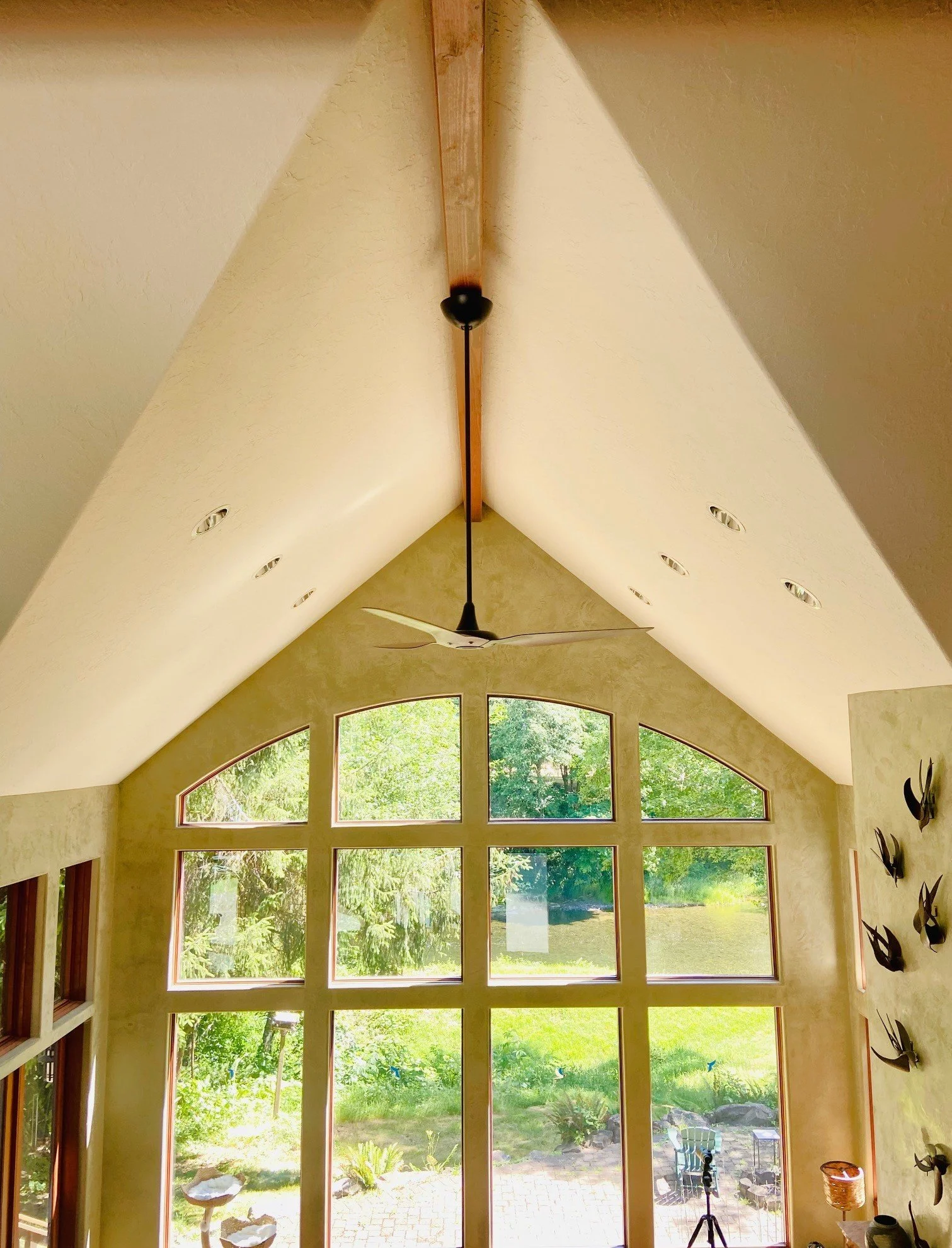Interior view of a room with a vaulted ceiling, large arched windows showing greenery outside, a ceiling fan, and wall-mounted decor.