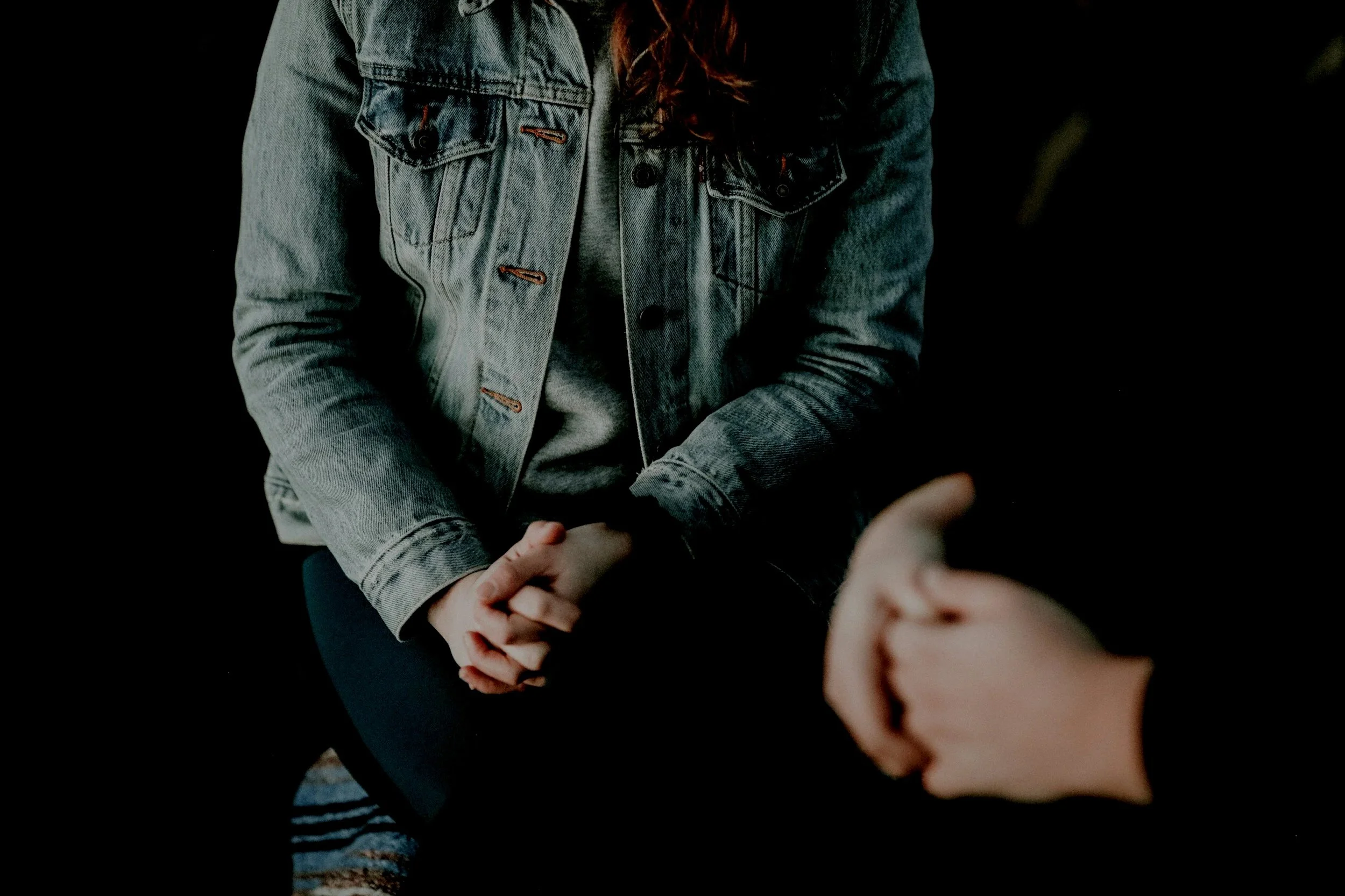 Person wearing a denim jacket and gray shirt, sitting with hands clasped, engaging in conversation with another person who has a thumb up.