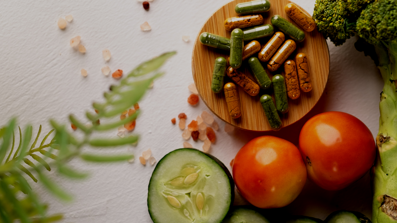 Fresh vegetables including a sliced cucumber, two tomatoes, and broccoli, along with a wooden dish filled with green and brown capsules, on a white surface with scattered Himalayan salt.