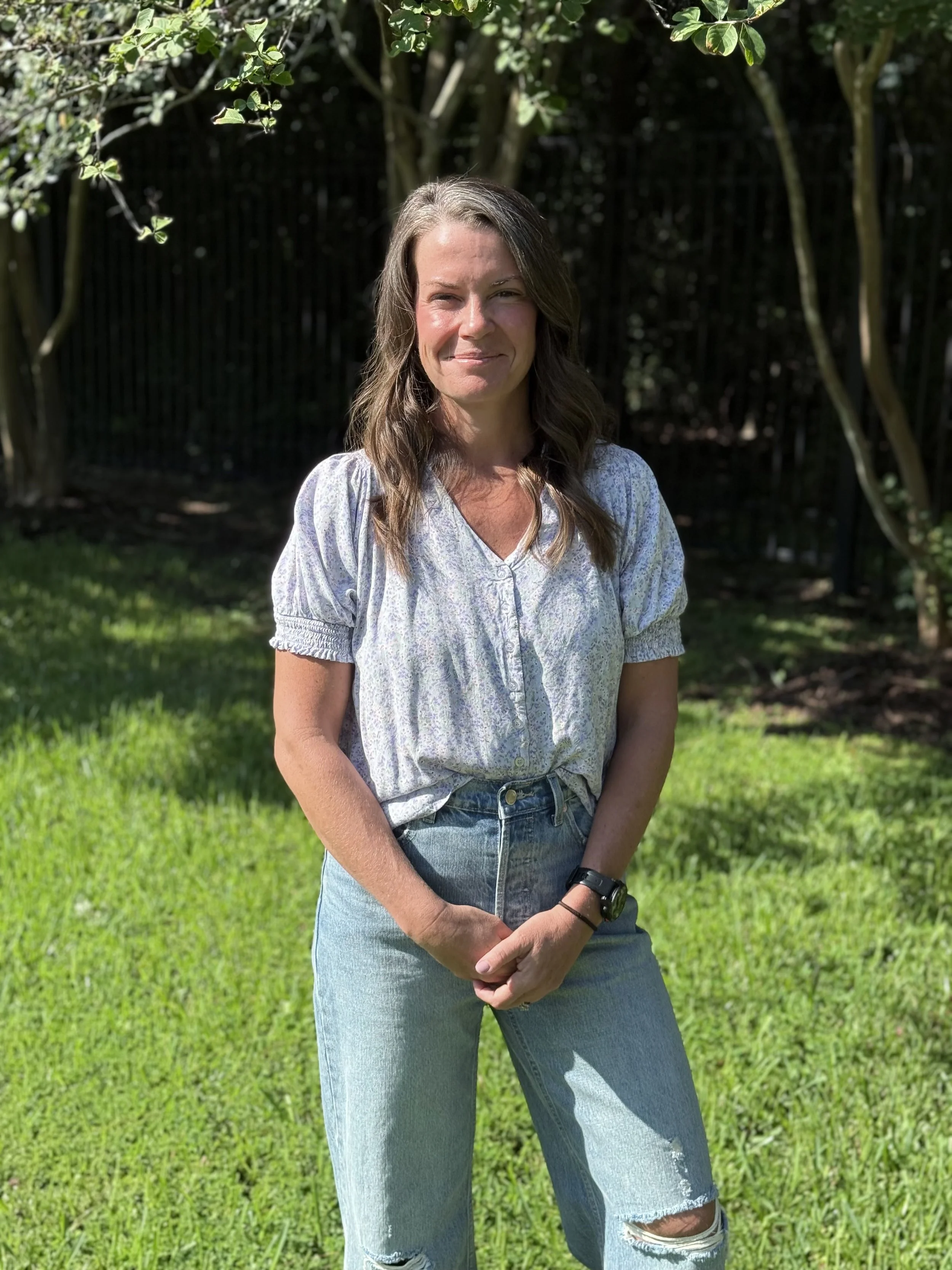 A woman standing outdoors on a grassy lawn, with trees and a black fence in the background. She is smiling slightly, wearing a light-colored short-sleeved blouse and ripped jeans, with a watch on her left wrist, and has wavy brown hair.