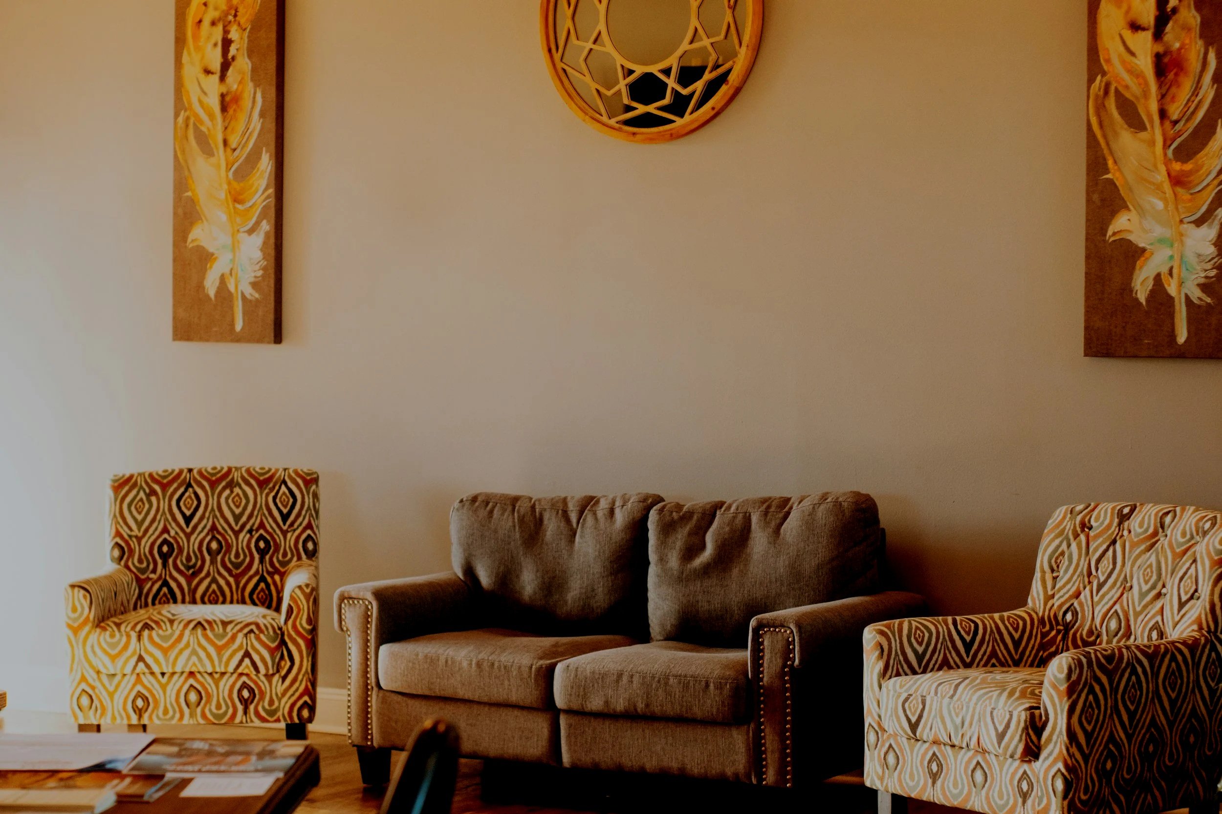 A living room with beige wall art featuring feathers, a round decorative wall piece, a beige sofa, and two armchairs with colorful geometric patterns.
