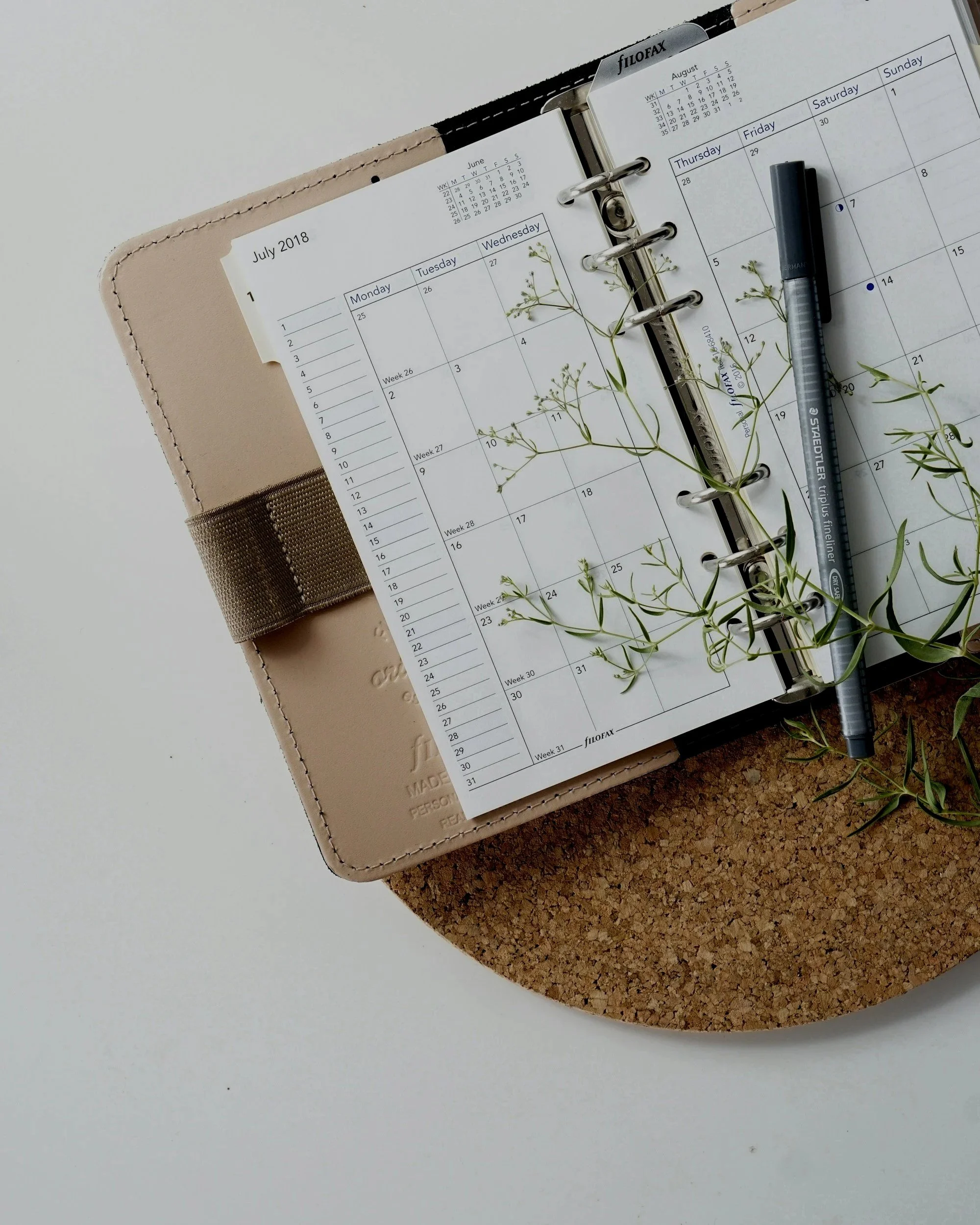 An open planner with July and August 2018 calendars, a gray pen, and green plant sprigs on a round cork mat, on a white surface.