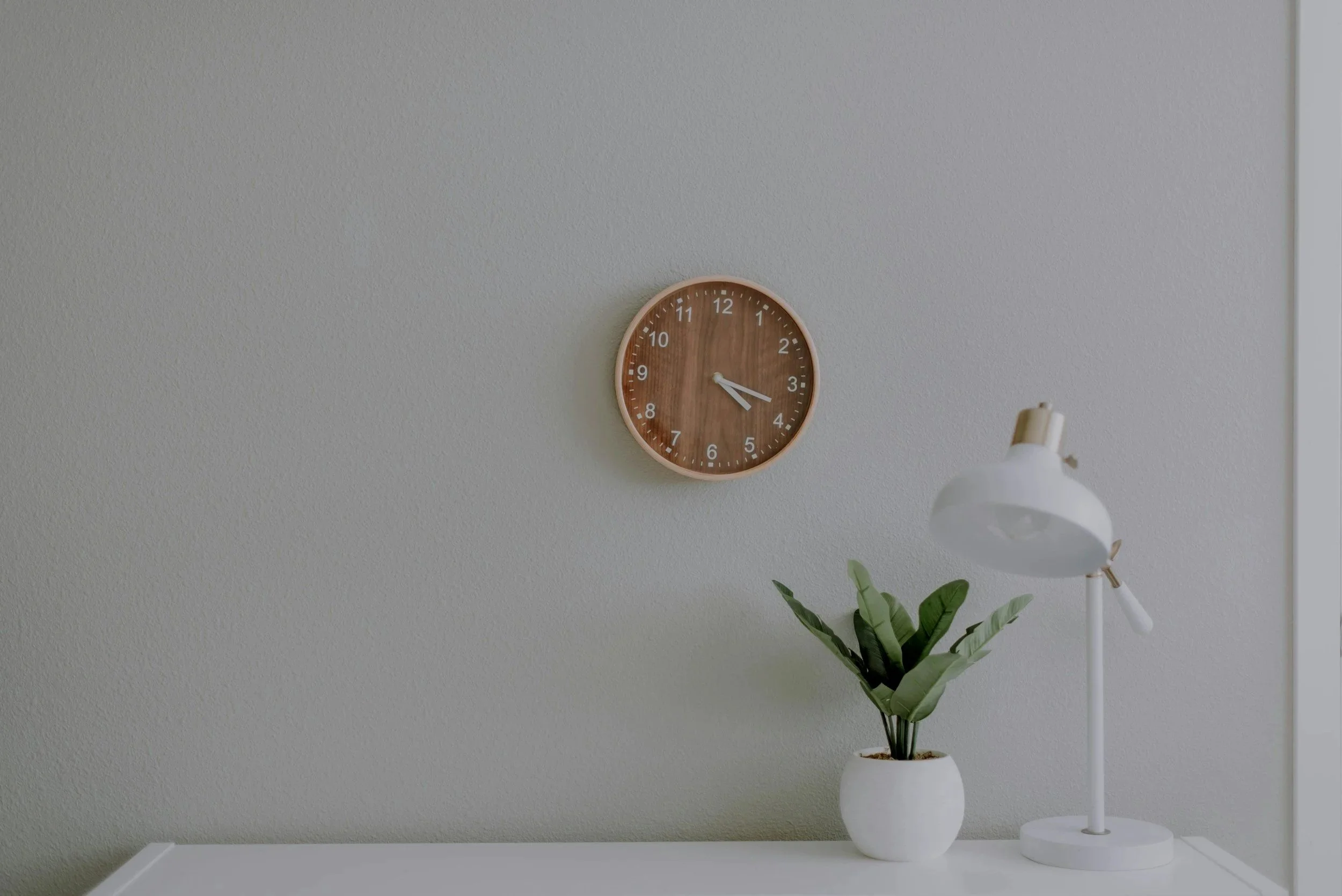 A wall clock showing approximately 3:20, a white desk lamp, and a potted green plant on a white desk against a plain textured gray wall.