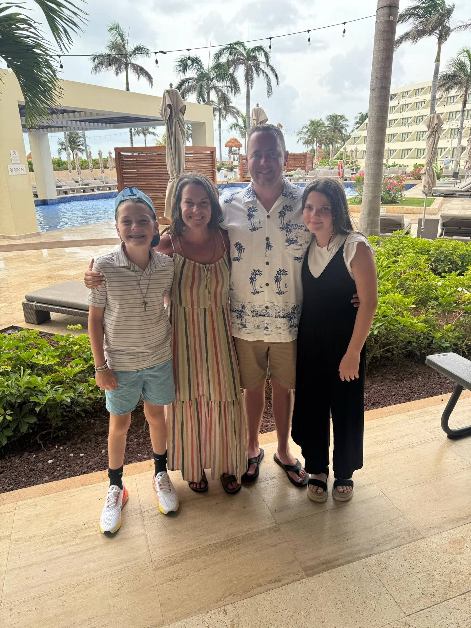 A family of four smiling, posing together at a poolside resort with palm trees, tropical landscape, and hotel buildings in the background.