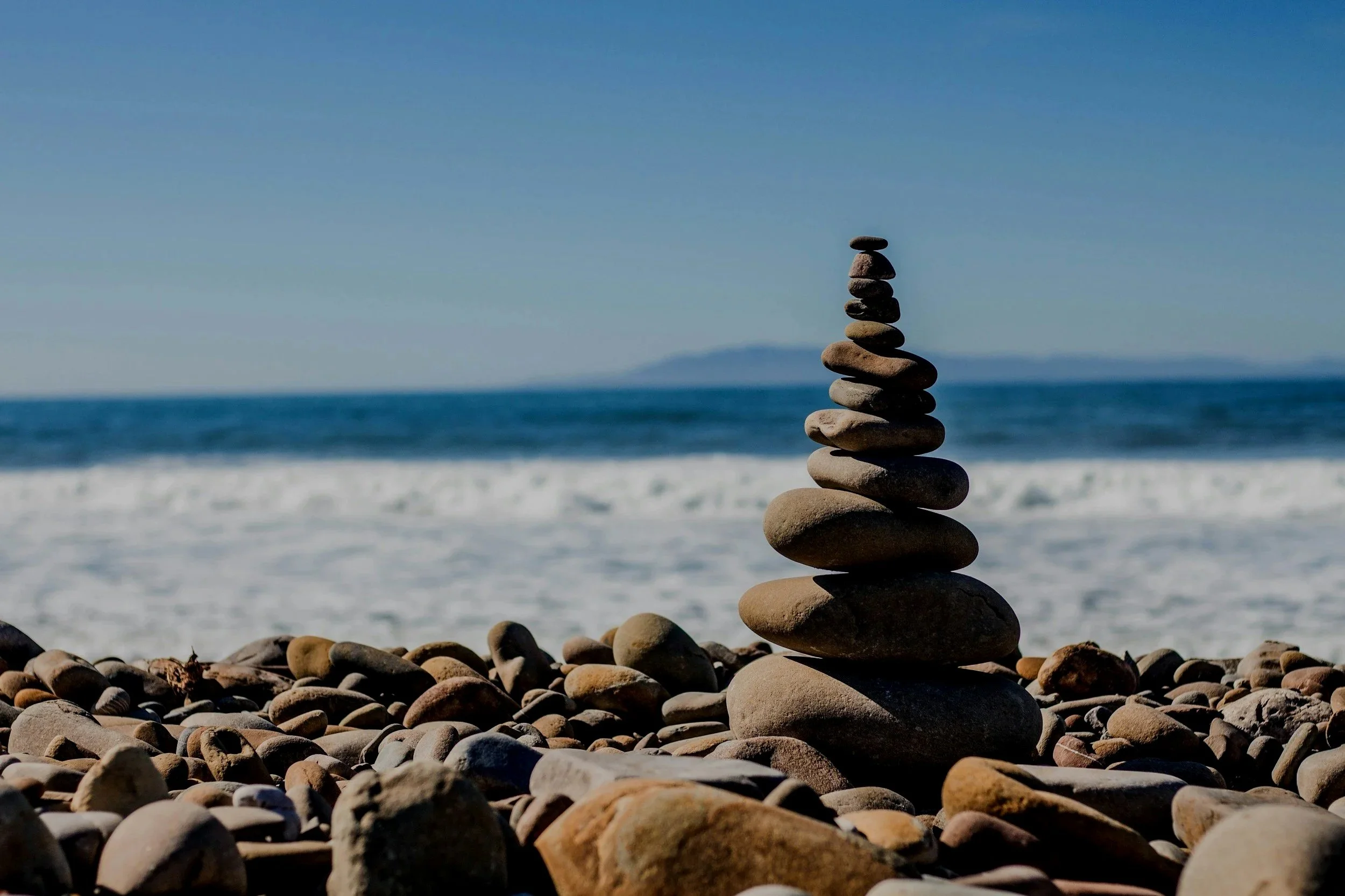 Stacked rocks on a pebble beach with ocean waves and a distant landmass under a clear blue sky.