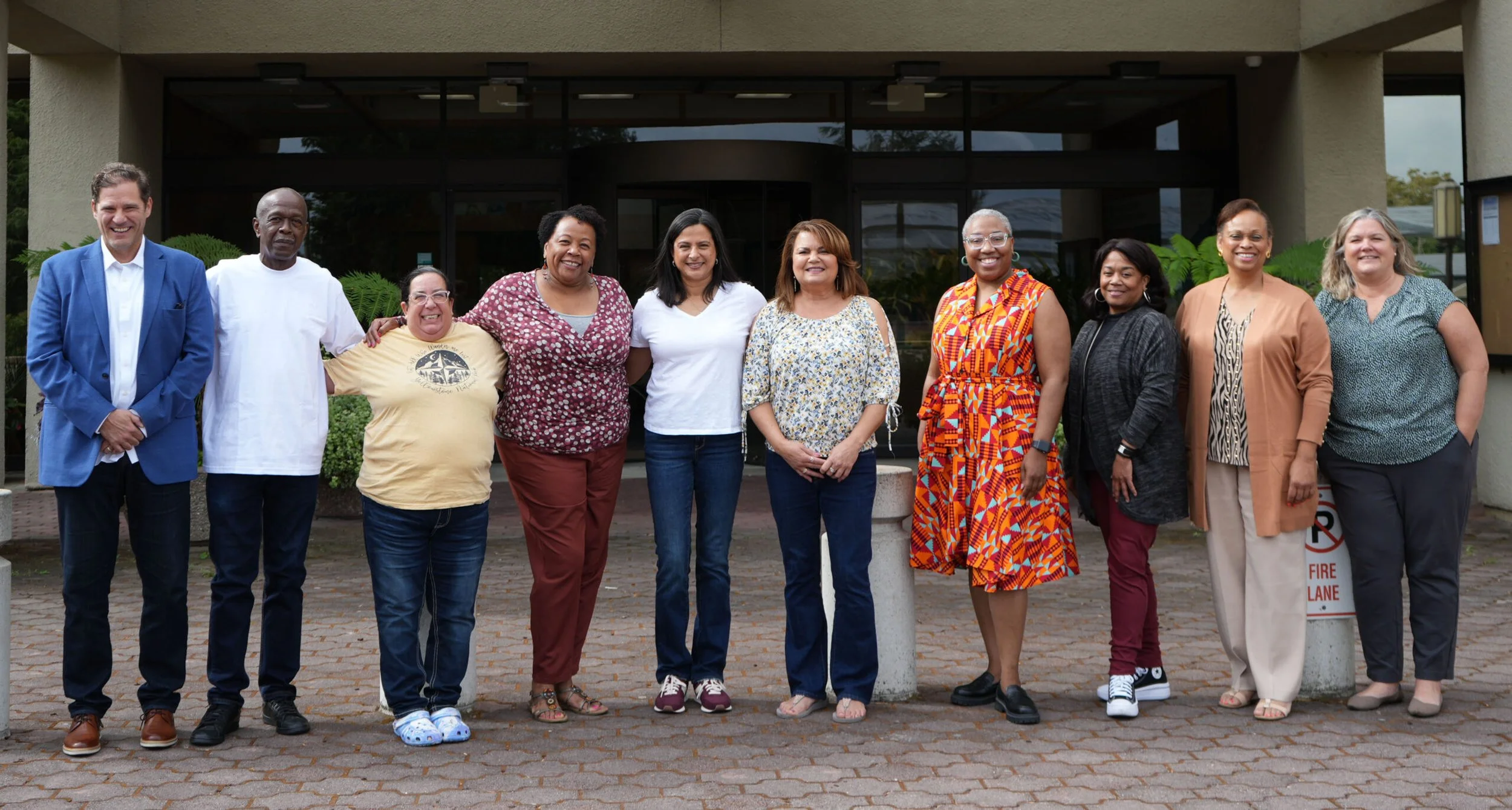 A diverse group of ten people representing the Community Advisory Council standing shoulder to shoulder outside a building, smiling for the photo.
