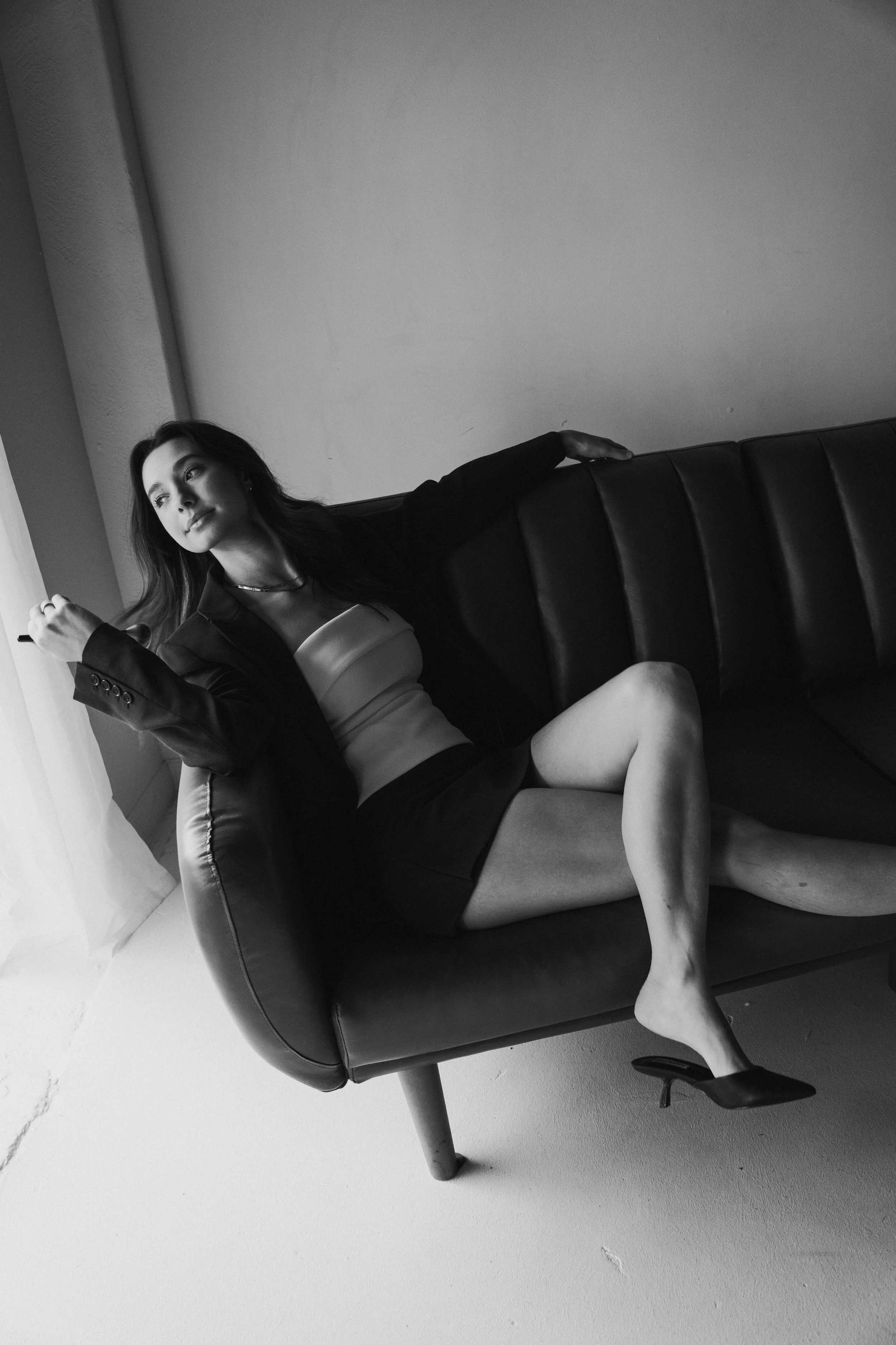 Black and white photo of a woman sitting on a couch near a window, with one leg stretched out and wearing high heels, a blazer, a top, and a skirt.