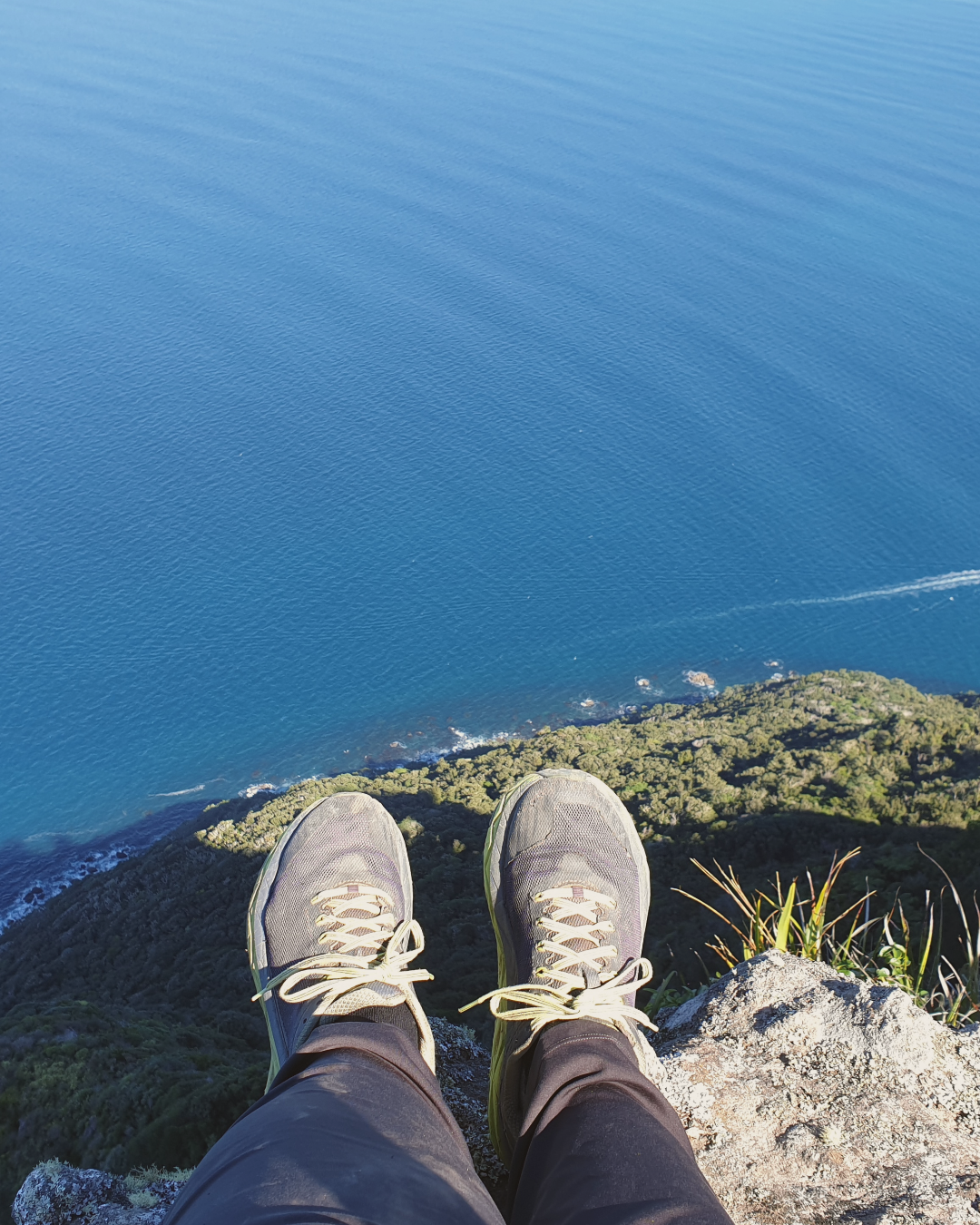 Feet hanging over the edge of a high cliff above deep blue water, symbolising the balance between sensitivity and intensity in dual-wired women.