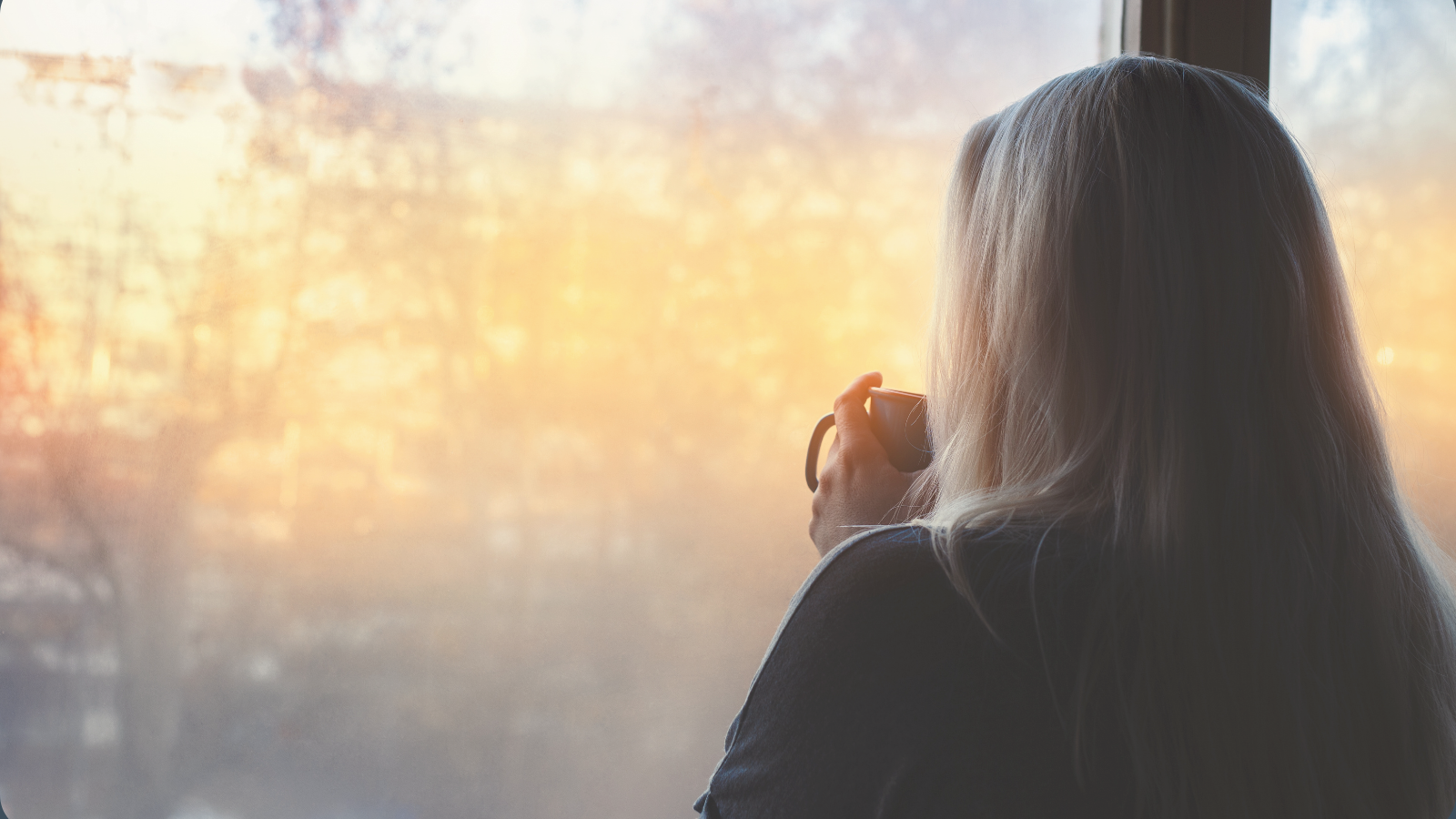 Woman holding a mug and looking out a window in quiet reflection, representing nervous system overwhelm, exhaustion, and the process of letting go.