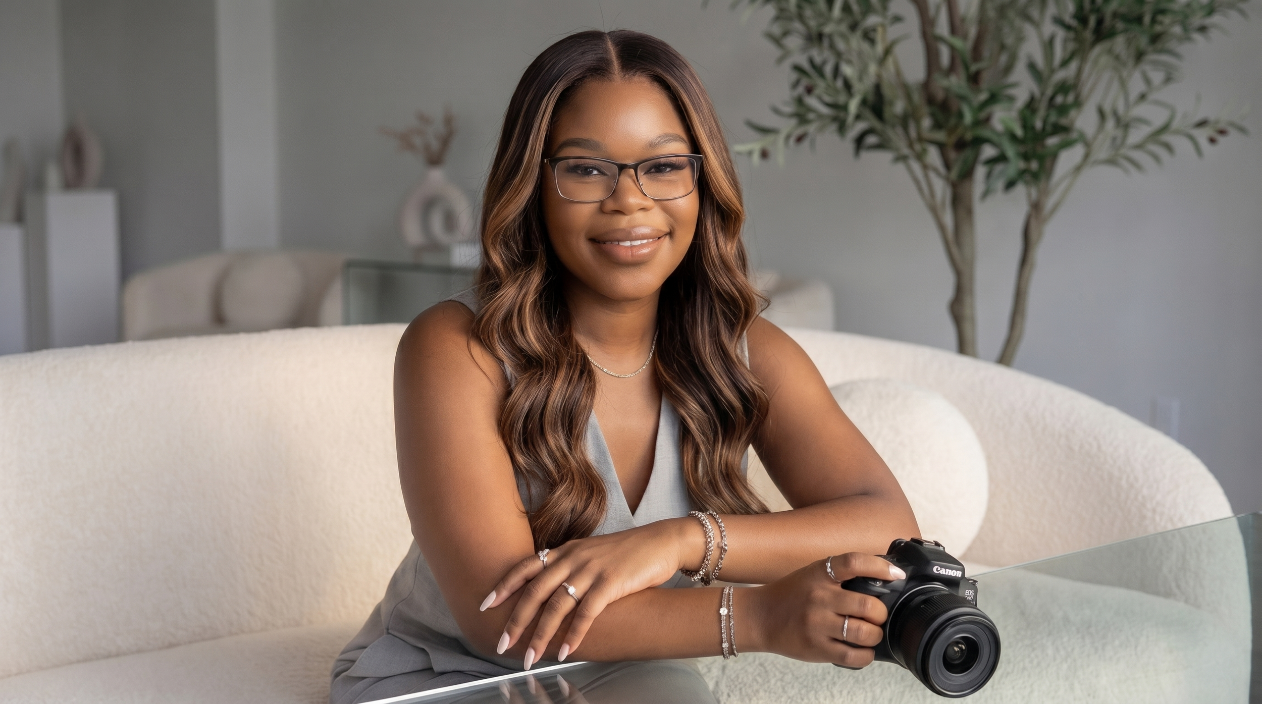 A young woman with long wavy hair, glasses, and jewelry sitting on a cream-colored couch holding a Canon camera, smiling in a modern living room with a large plant in the background.