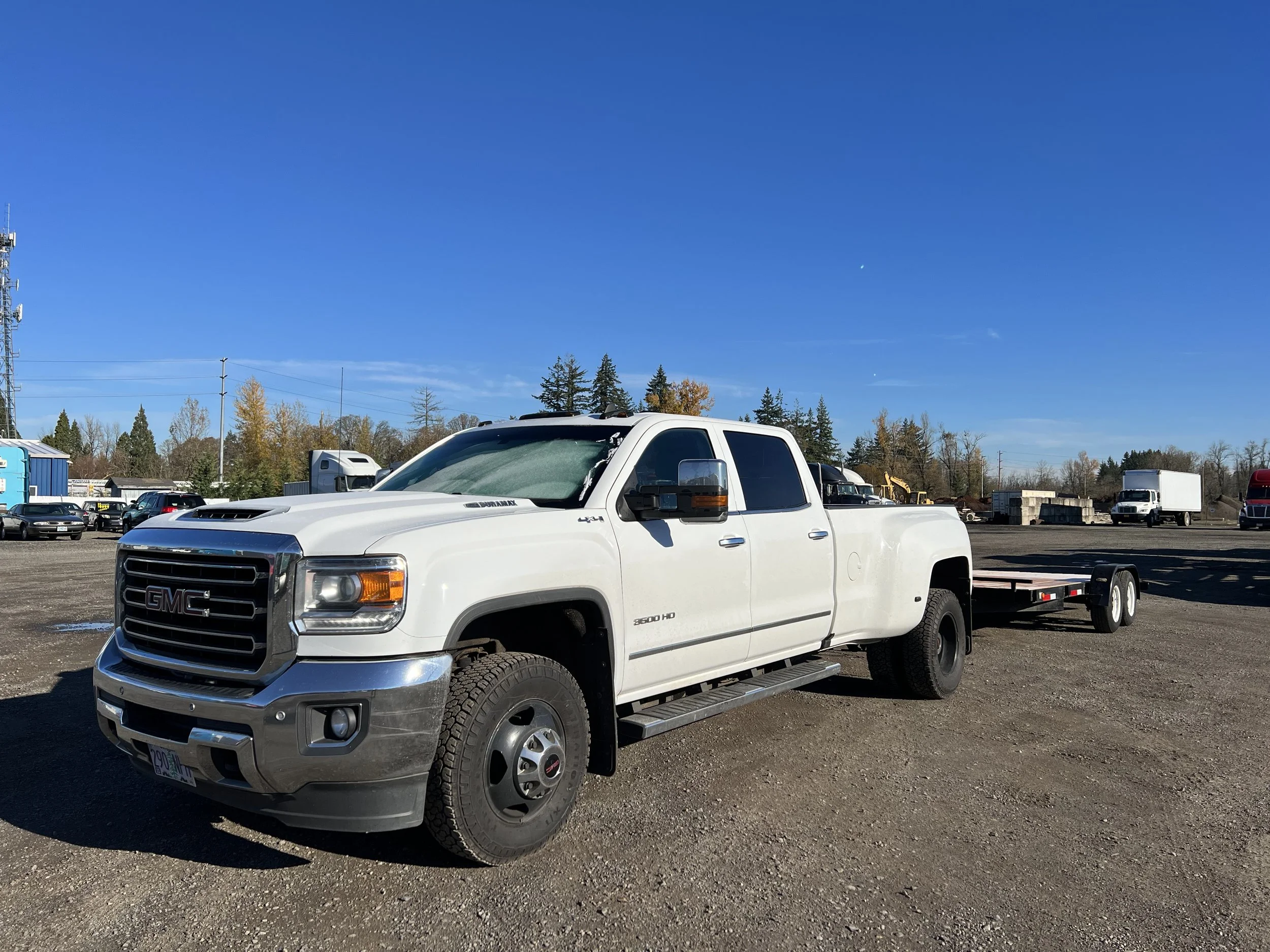 White GMC pickup truck with a flatbed trailer parked on a gravel lot, with trees and other vehicles in the background under a clear blue sky.