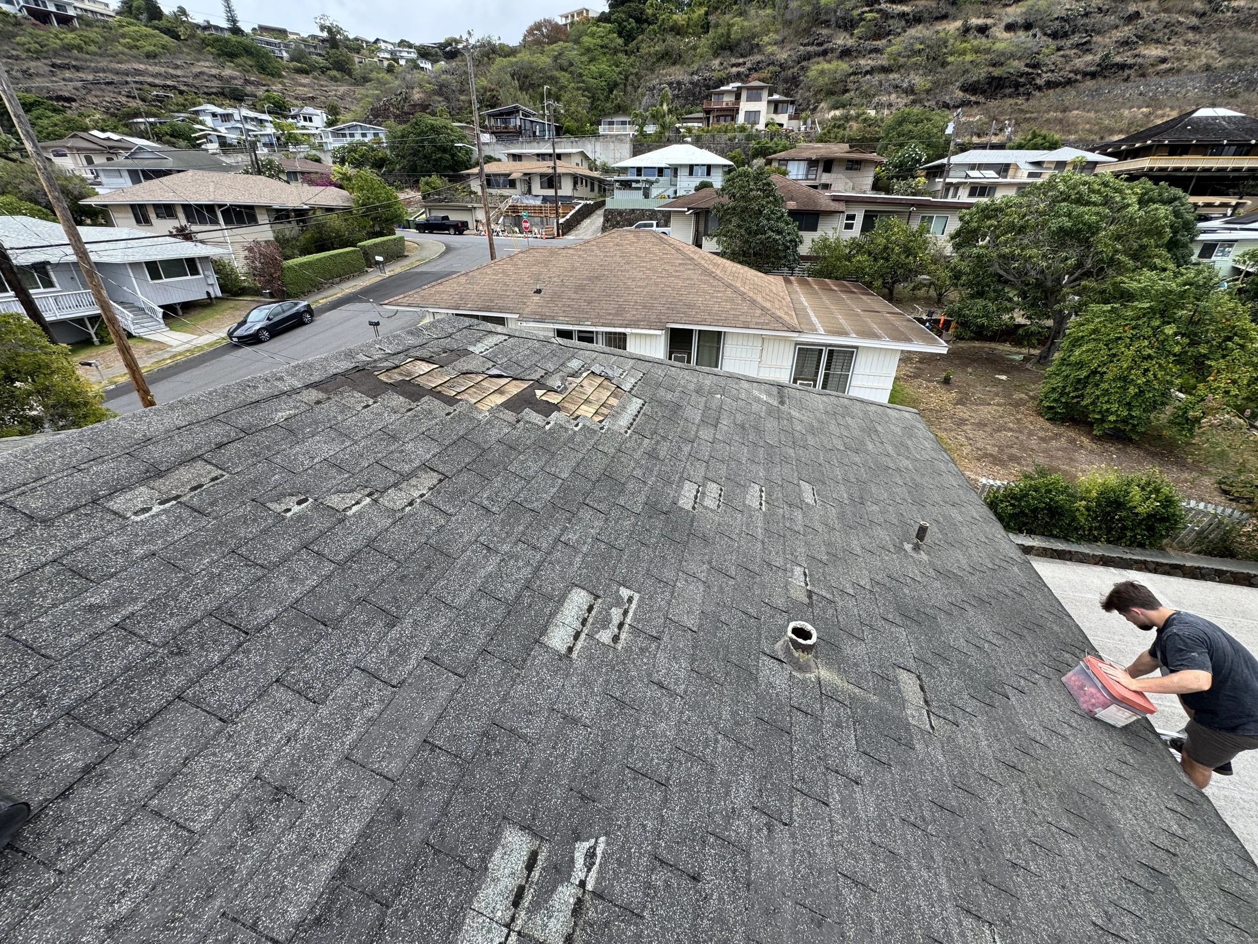 Person standing on a sloped roof, sorting through a plastic container, with a neighborhood of houses and trees in the background.