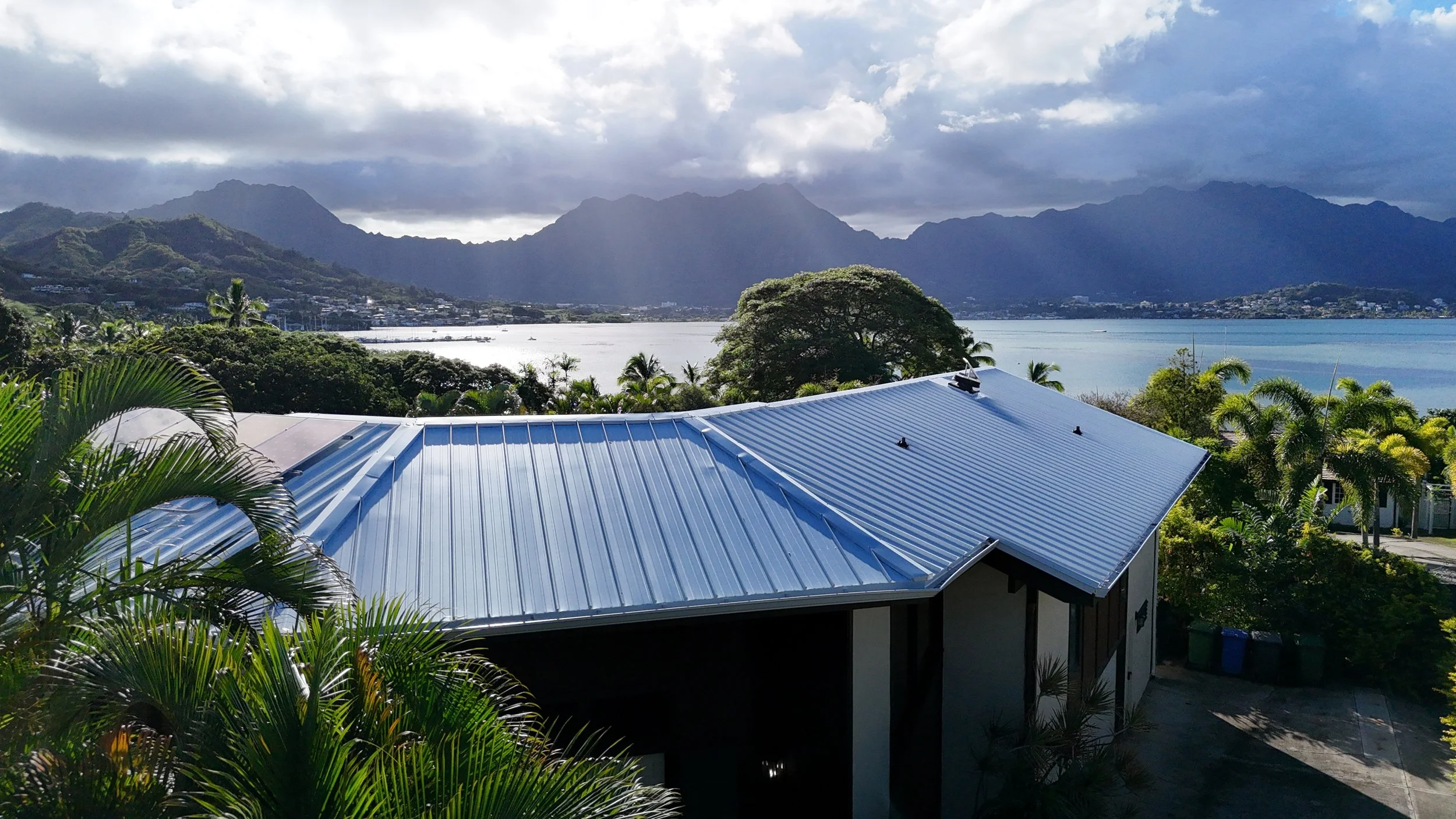 A house with a blue metal roof, surrounded by lush tropical trees and plants, overlooking a body of water and mountains in the distance under a cloudy sky.