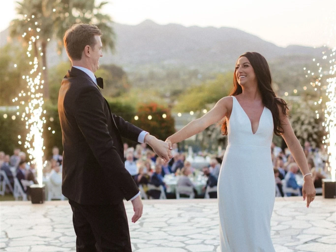 A bride and groom dancing at their outdoor wedding reception during sunset, with guests seated at tables in the background and fireworks or sparklers nearby.