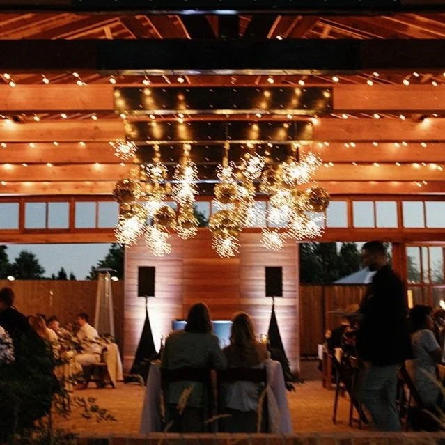 Indoor event space with wooden walls and ceiling, decorated with hanging lights and sparkler-like arrangements. Several groups of people seated at tables, some watching a presentation at a table with two women and a man standing nearby.