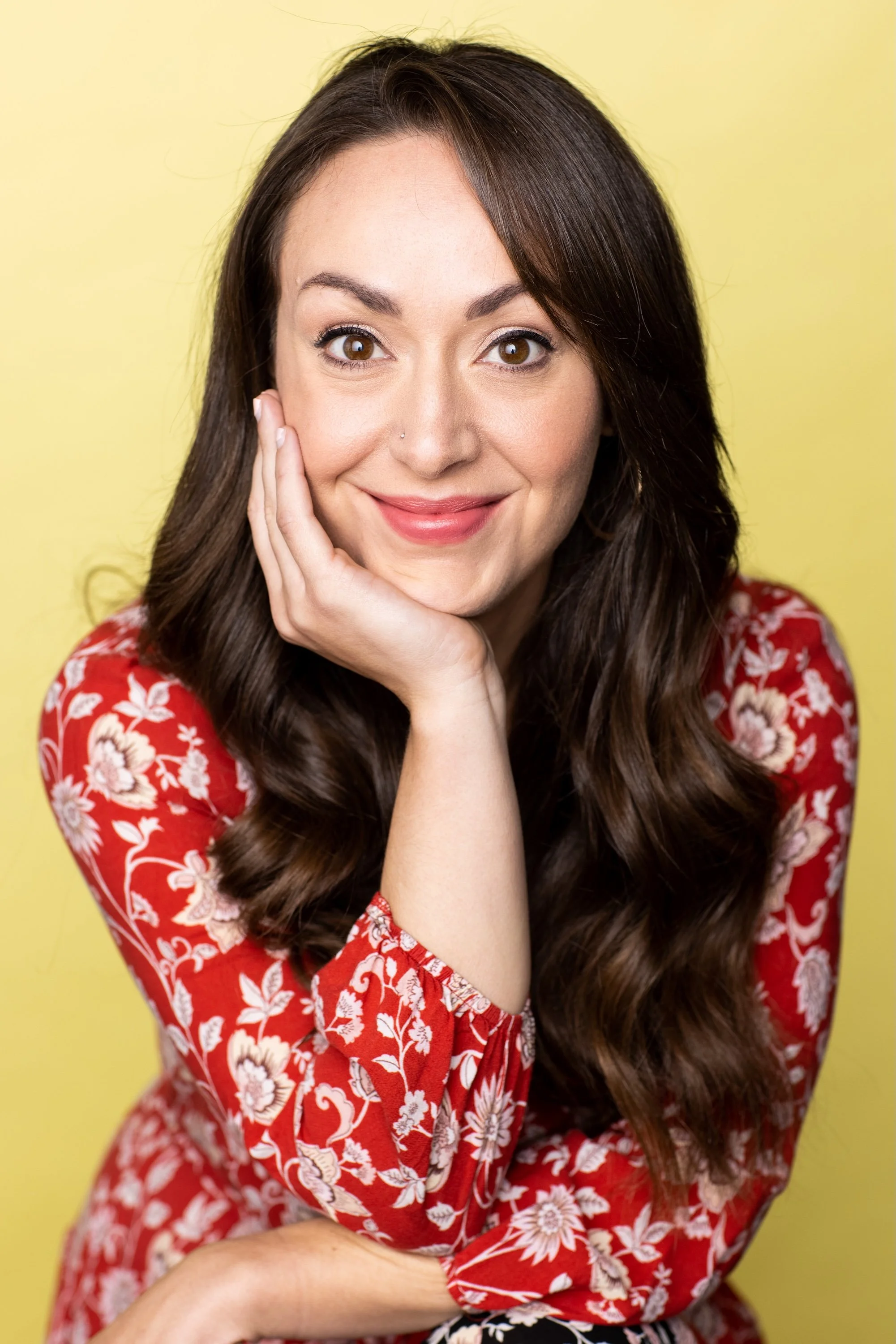 A woman with long brown hair, wearing a red floral top, smiling with her hand resting on her chin against a yellow background.