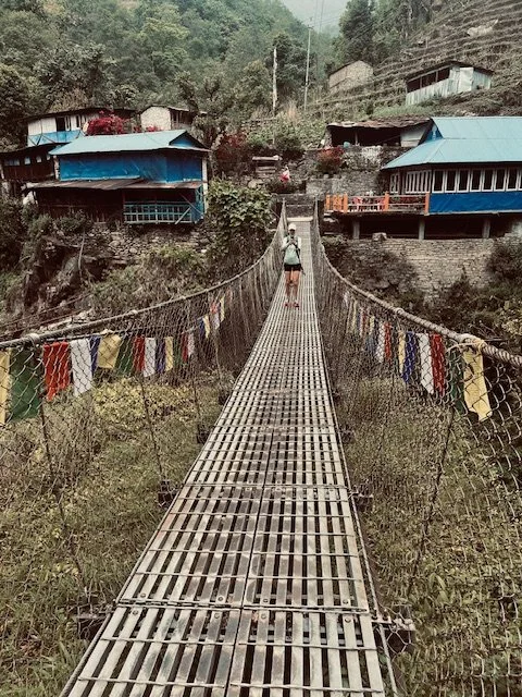 Person walking on a suspension bridge with colorful prayer flags, over a valley with houses and trees on hillsides.