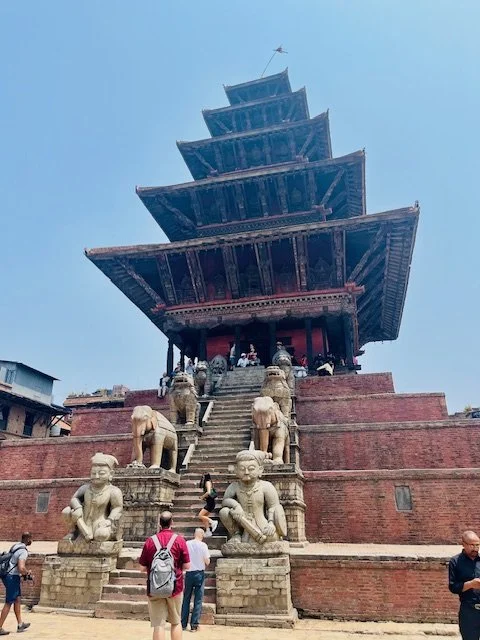 A traditional multi-tiered pagoda-style temple with stairs flanked by elephant statues and guardian figures in the foreground, set against a blue sky.