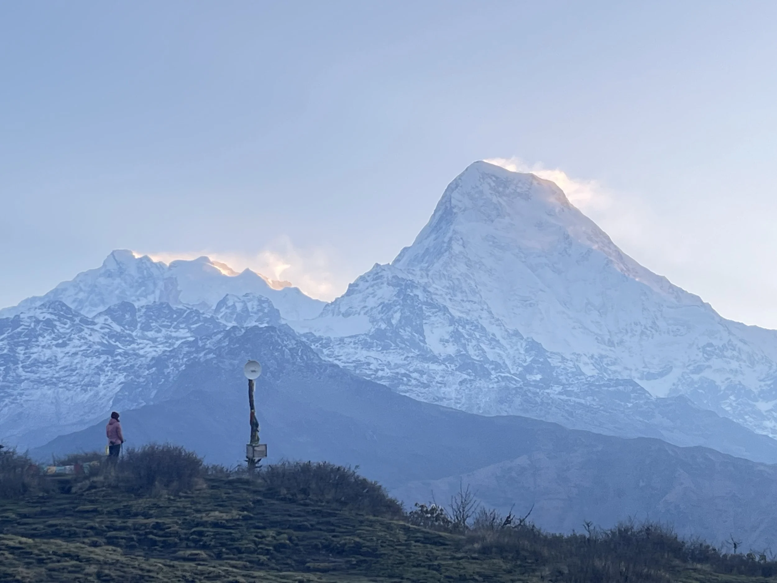A person standing on a grassy hill with snow-capped mountains in the background, and a lamp post nearby.