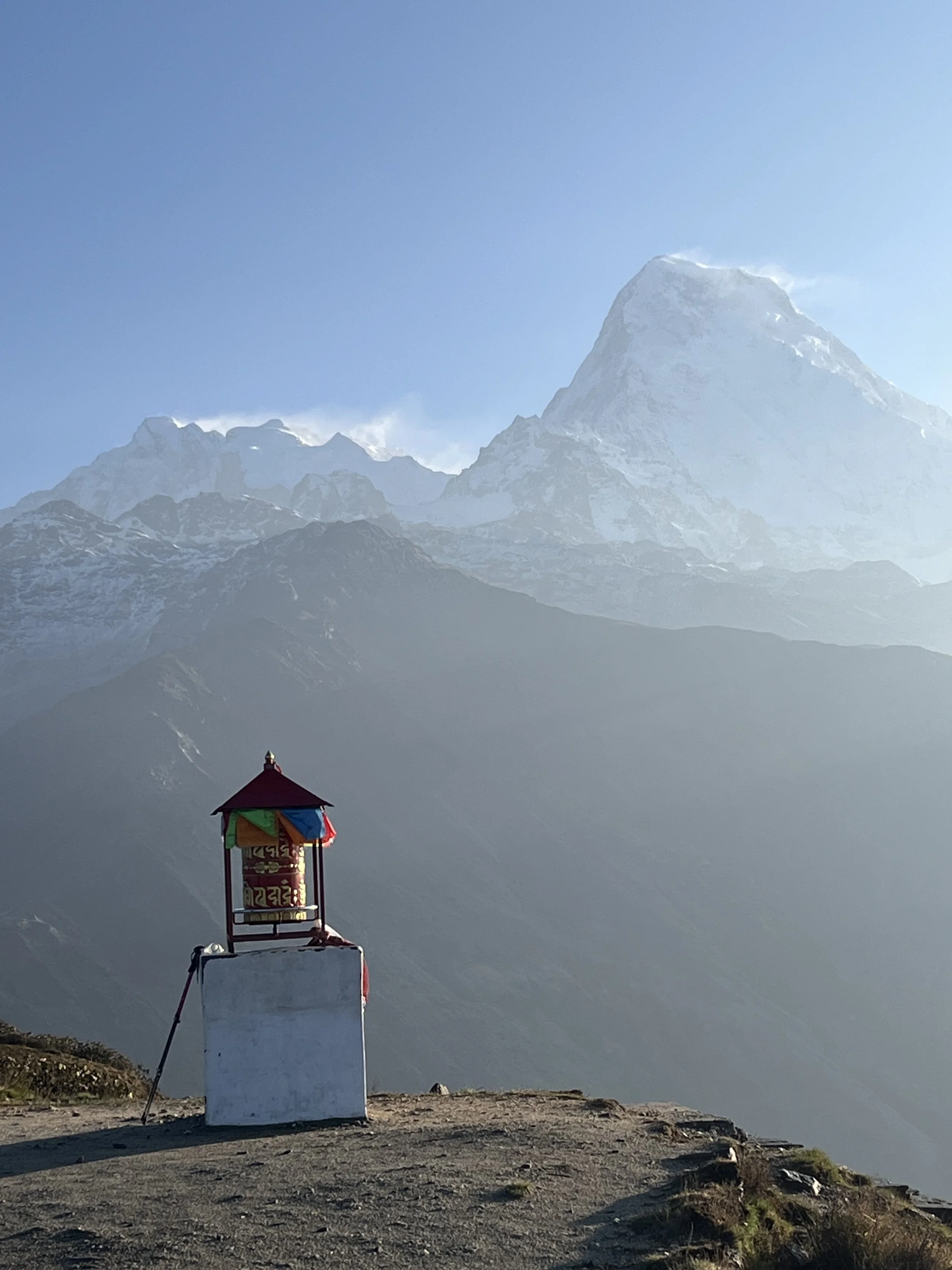 A small shrine with colorful prayer flags on a white base, set against snowy mountains and a clear blue sky.