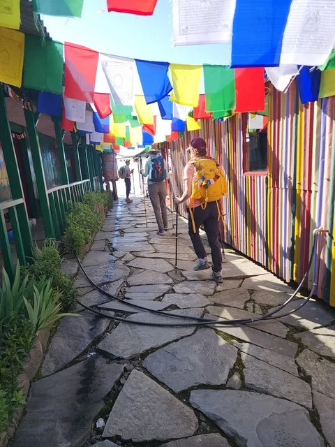 People walking along a stone pathway with colorful prayer flags hanging overhead.
